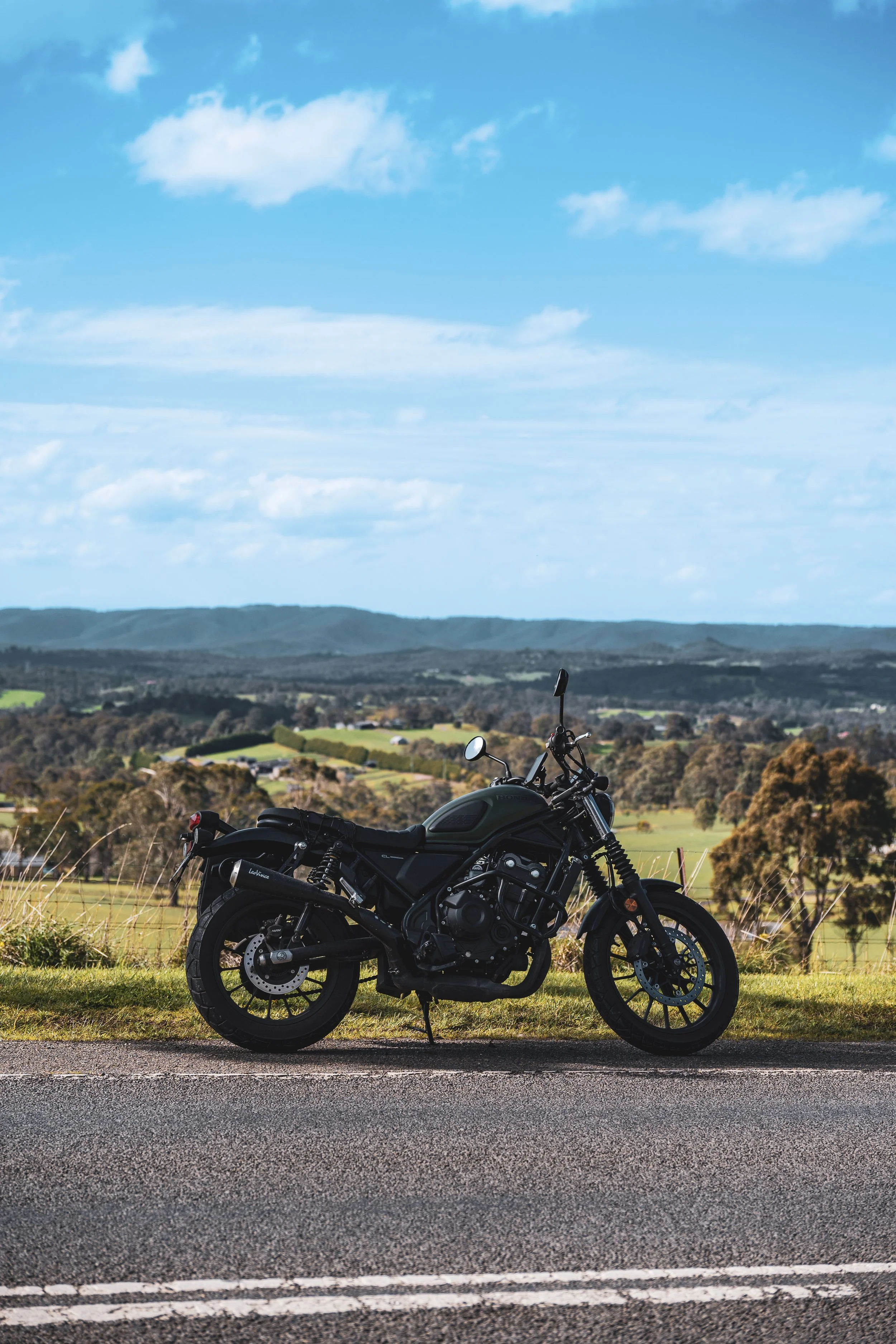 A black motorcycle parked on the side of a rural road with rolling fields, trees, and hills in the background under a partly cloudy blue sky.