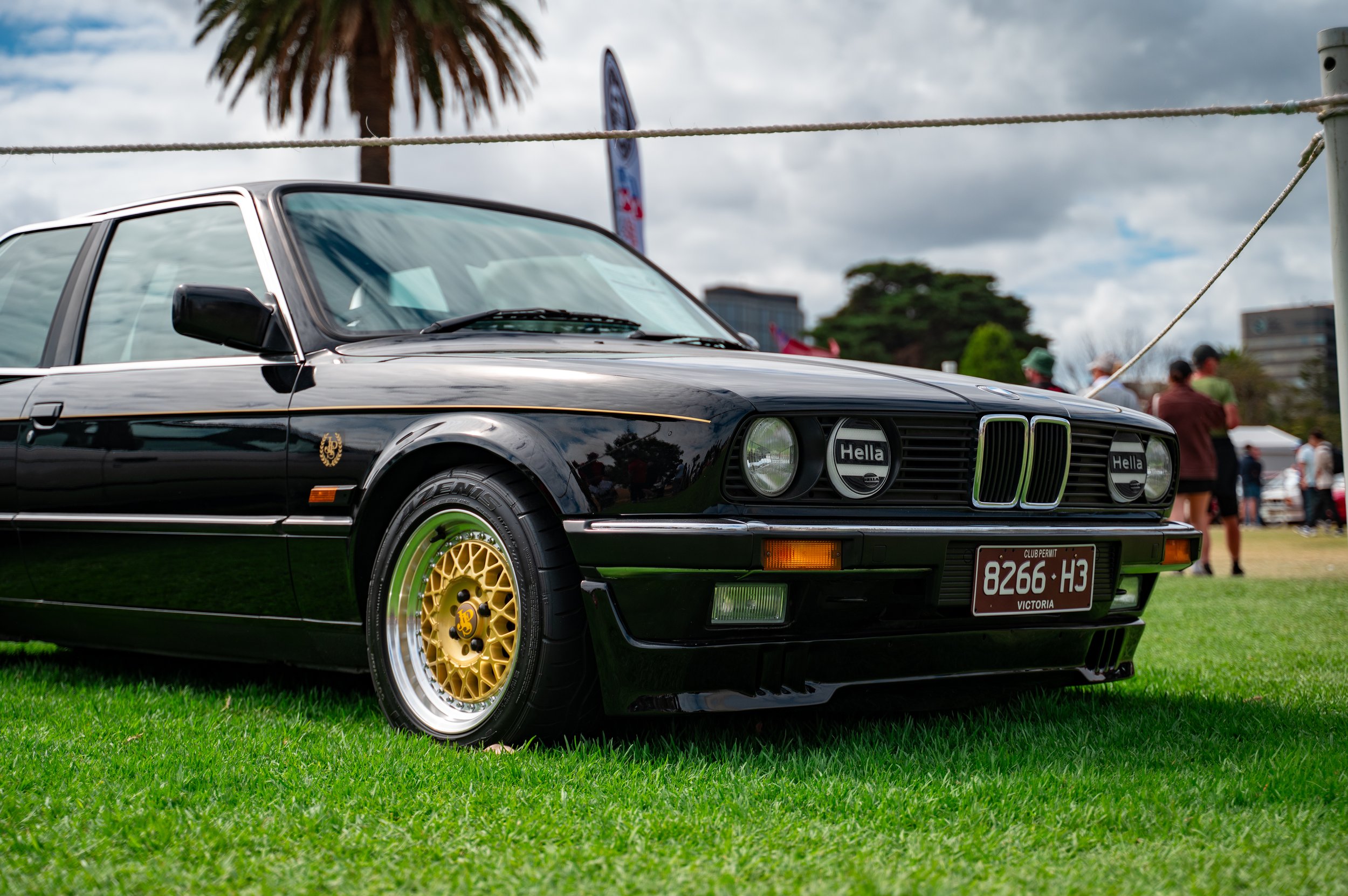 A vintage black BMW car with gold wheels on grass at a car show, with people and palm trees in the background.