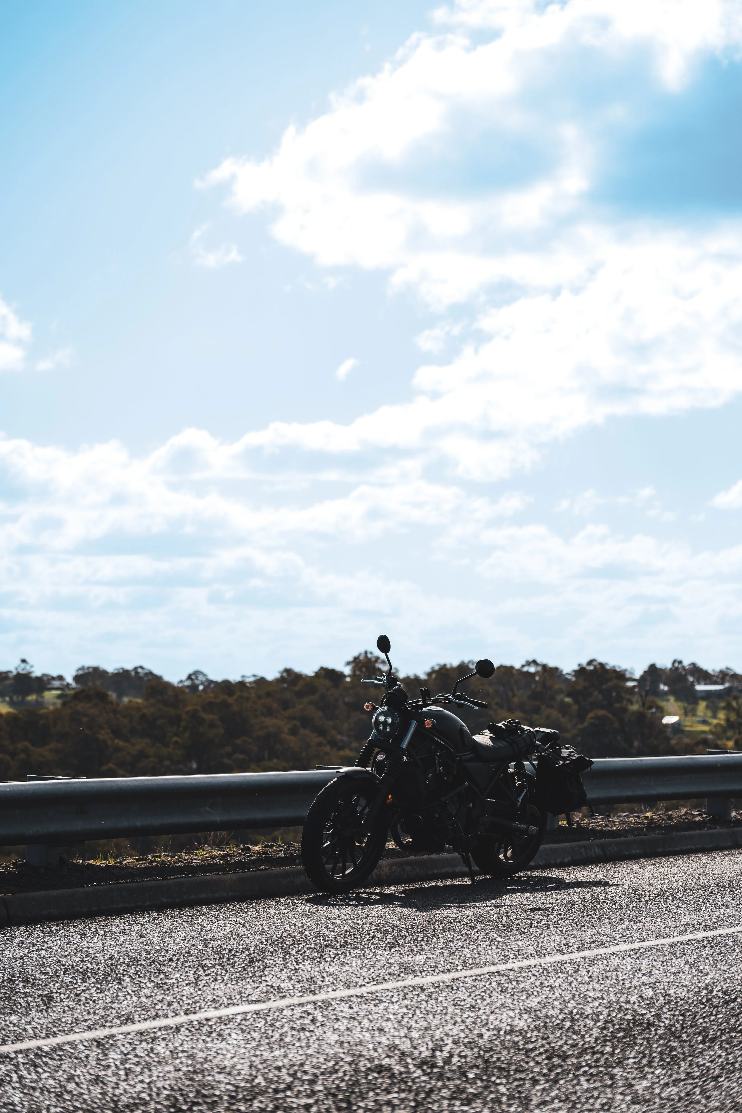 A black motorcycle parked on the side of a paved road with a metal guardrail, with a backdrop of green trees and a cloudy sky.