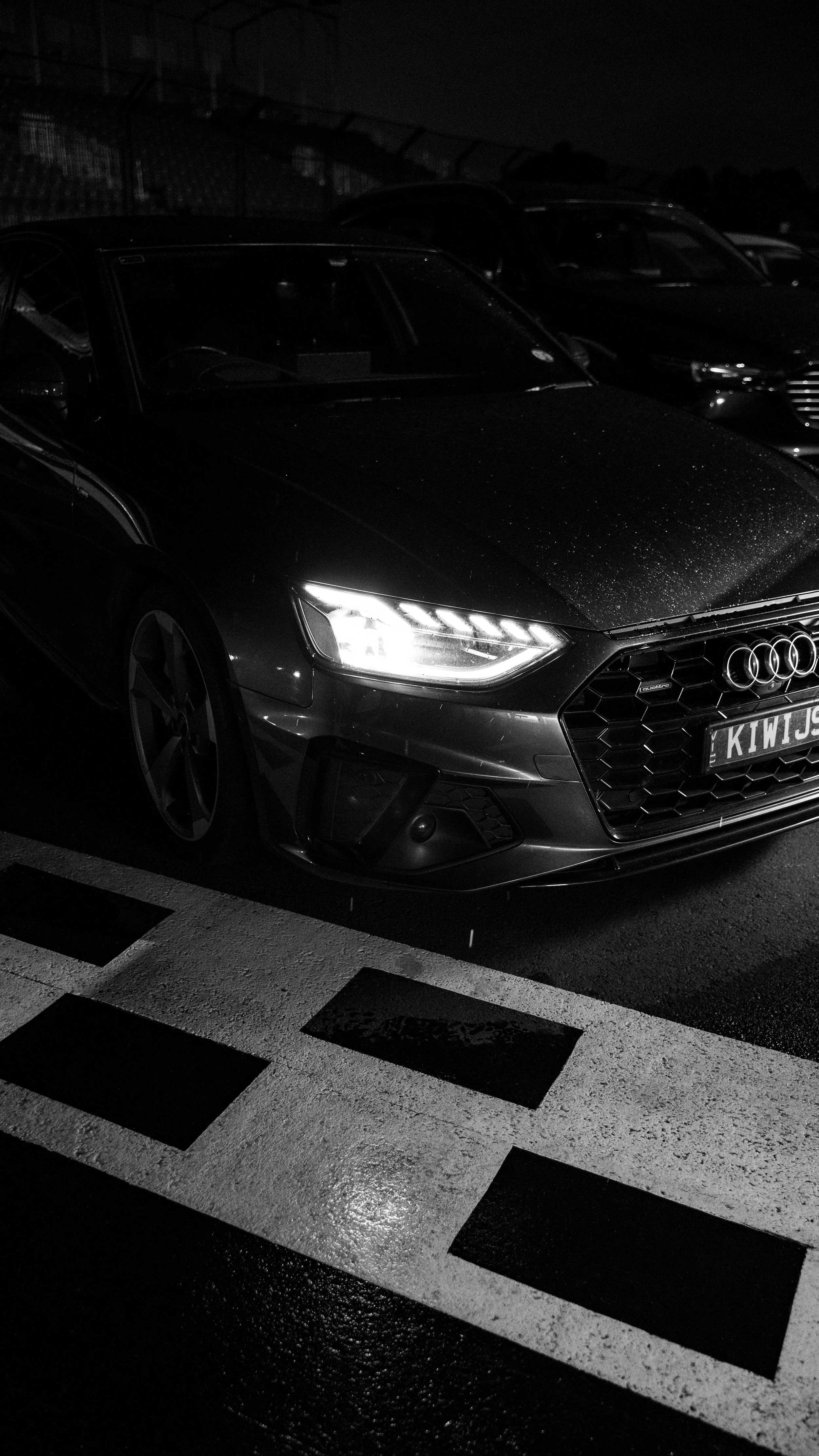 A dark-colored Audi car parked in a parking lot at night, with its headlights on and a pickup truck visible in the background.