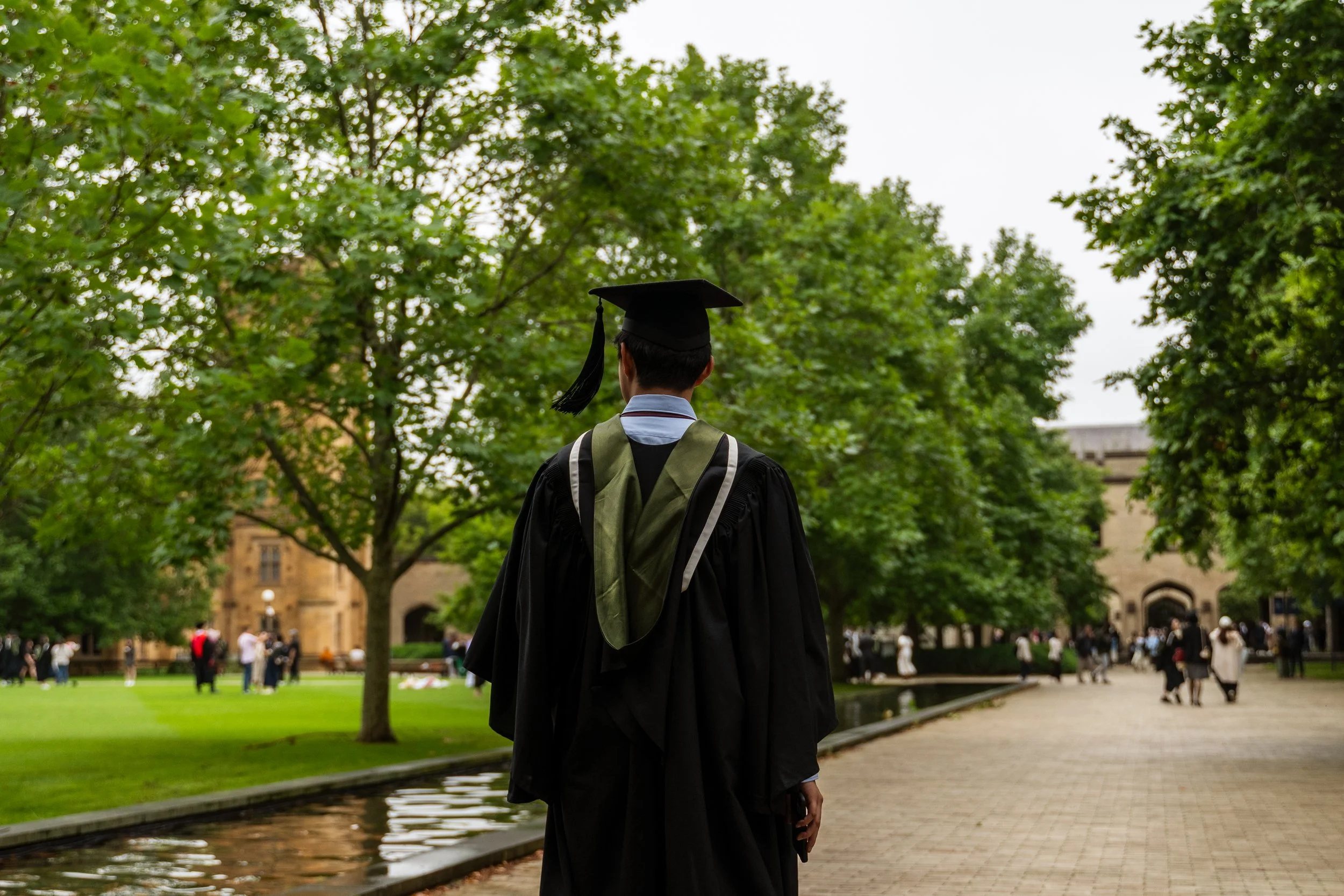 A graduate in a cap and gown walking outdoors on a university campus with trees and other graduates and people in the background.