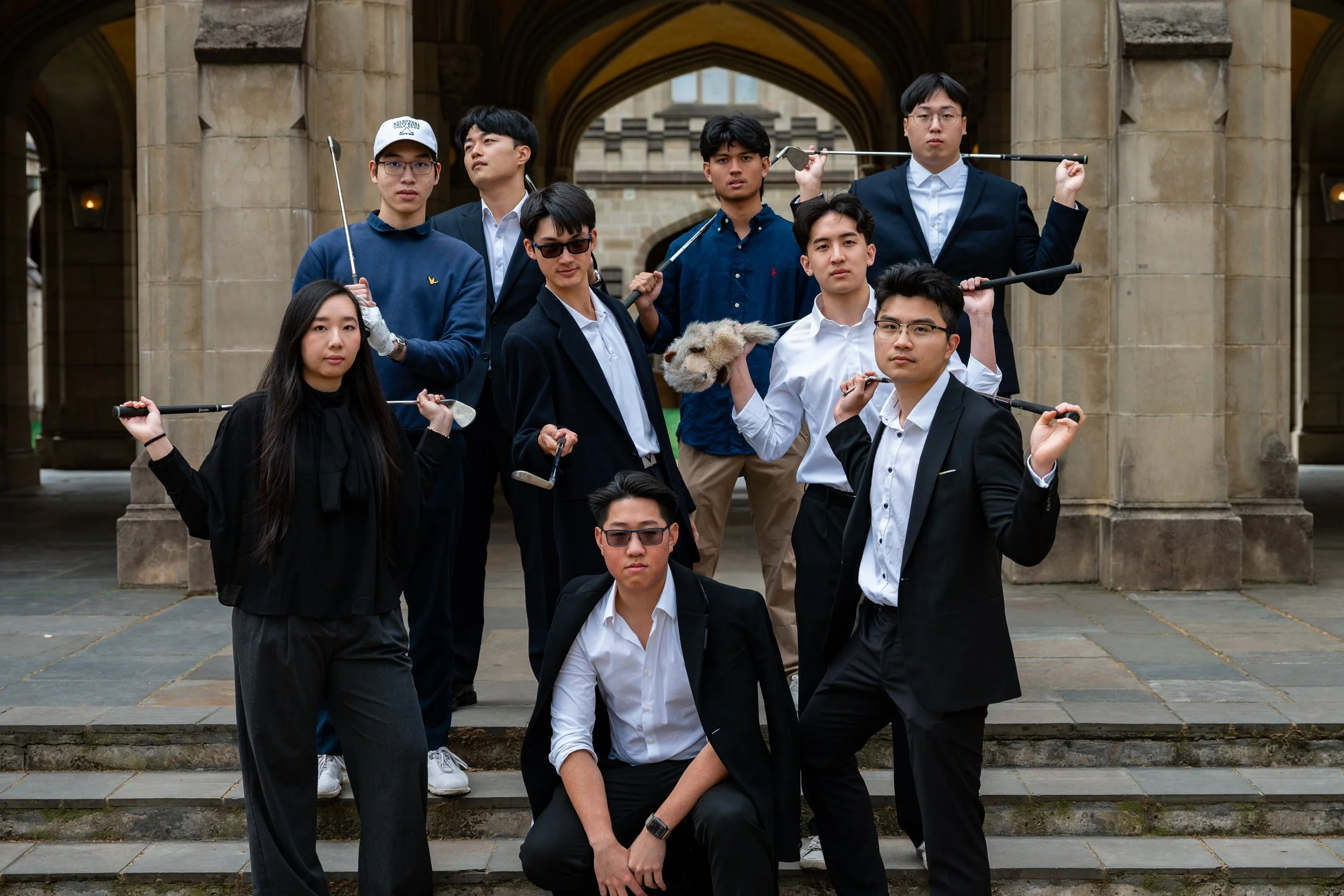 A group of young adults dressed in formal and semi-formal attire posing outdoors on stone steps, holding golf clubs and a stuffed animal.
