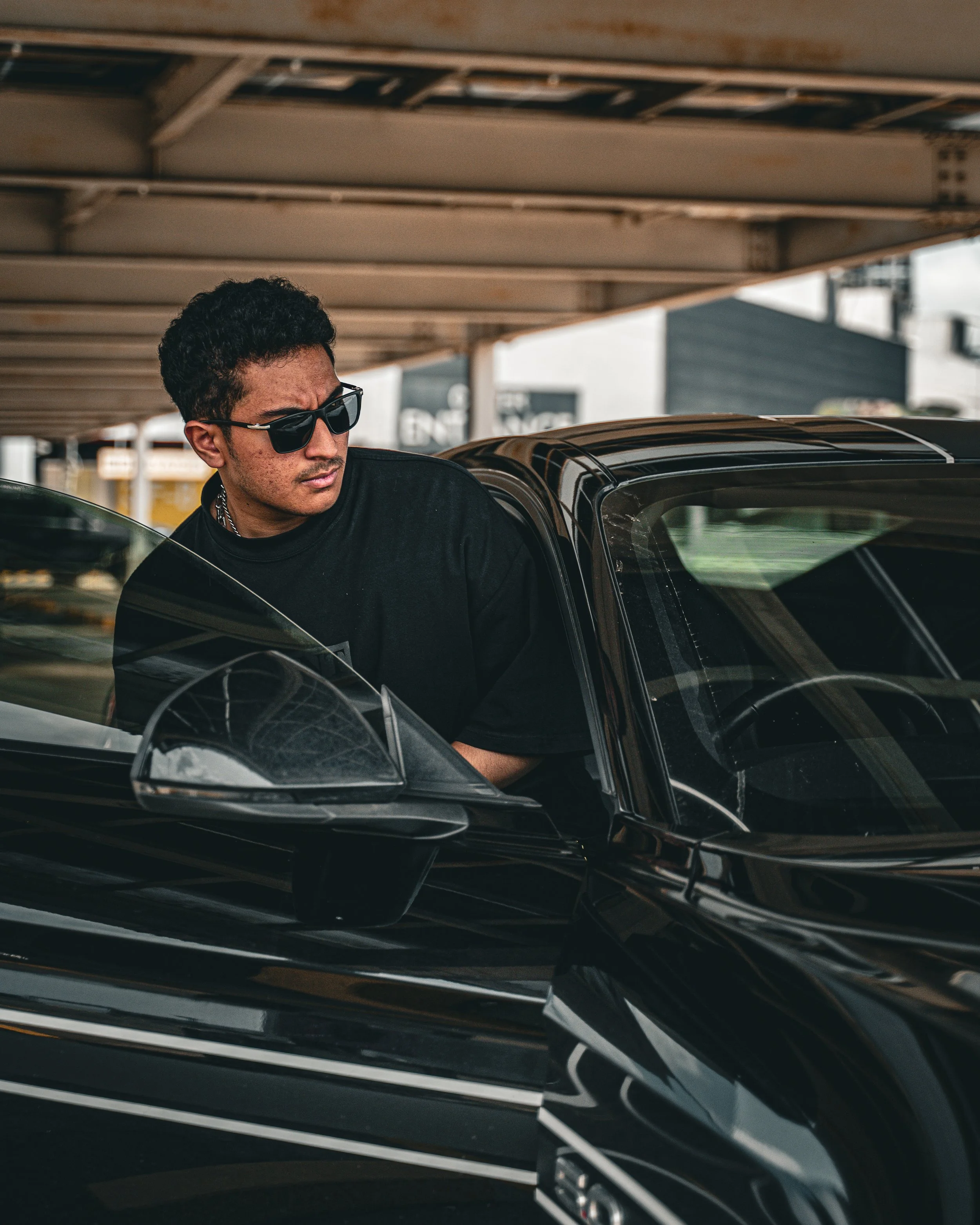 A young man with curly hair wearing sunglasses and a black shirt, leaning into the open door of a sleek black car, under a parking structure.
