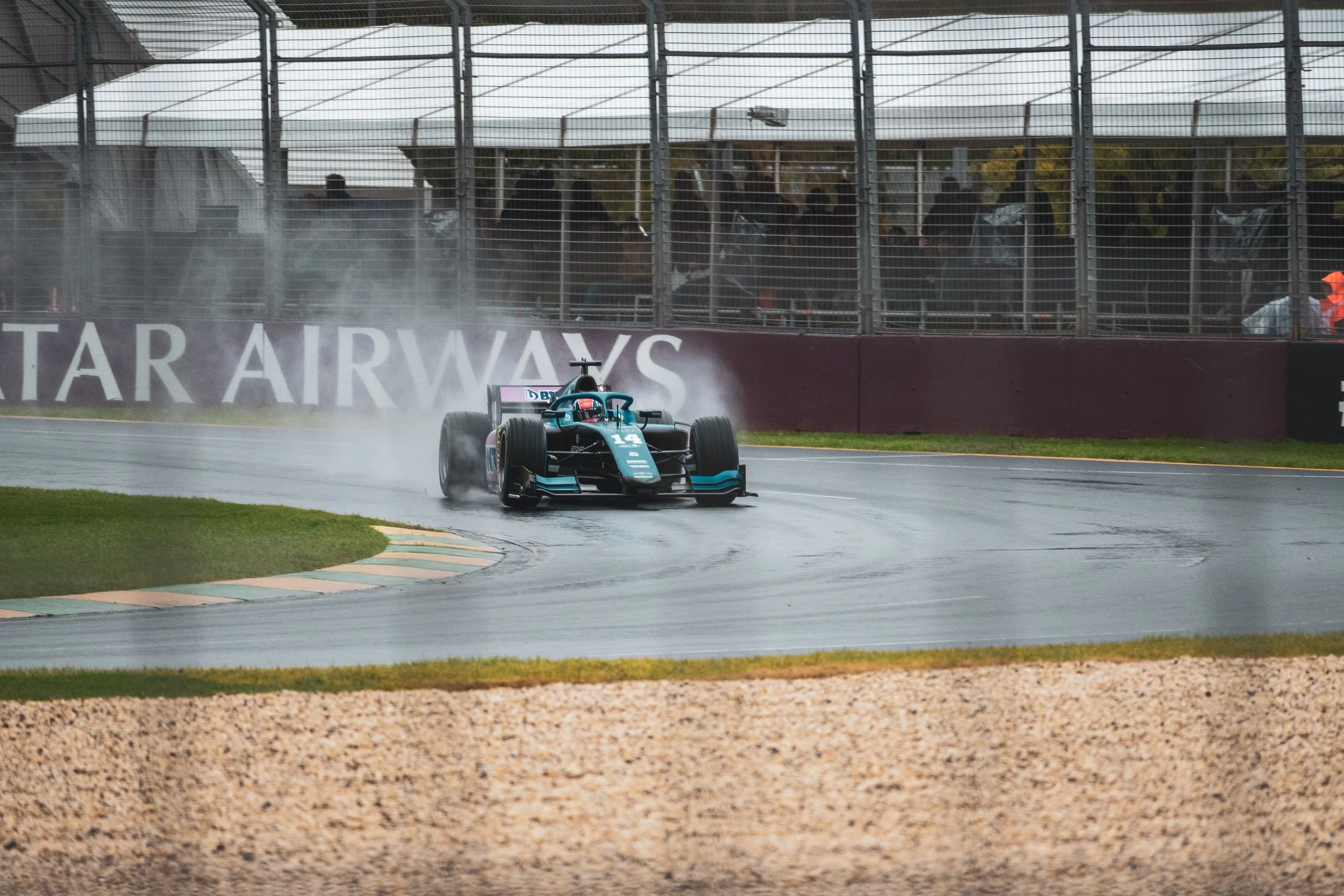 Race car on wet track during rain at a motorsport event, with spectators behind a fence.