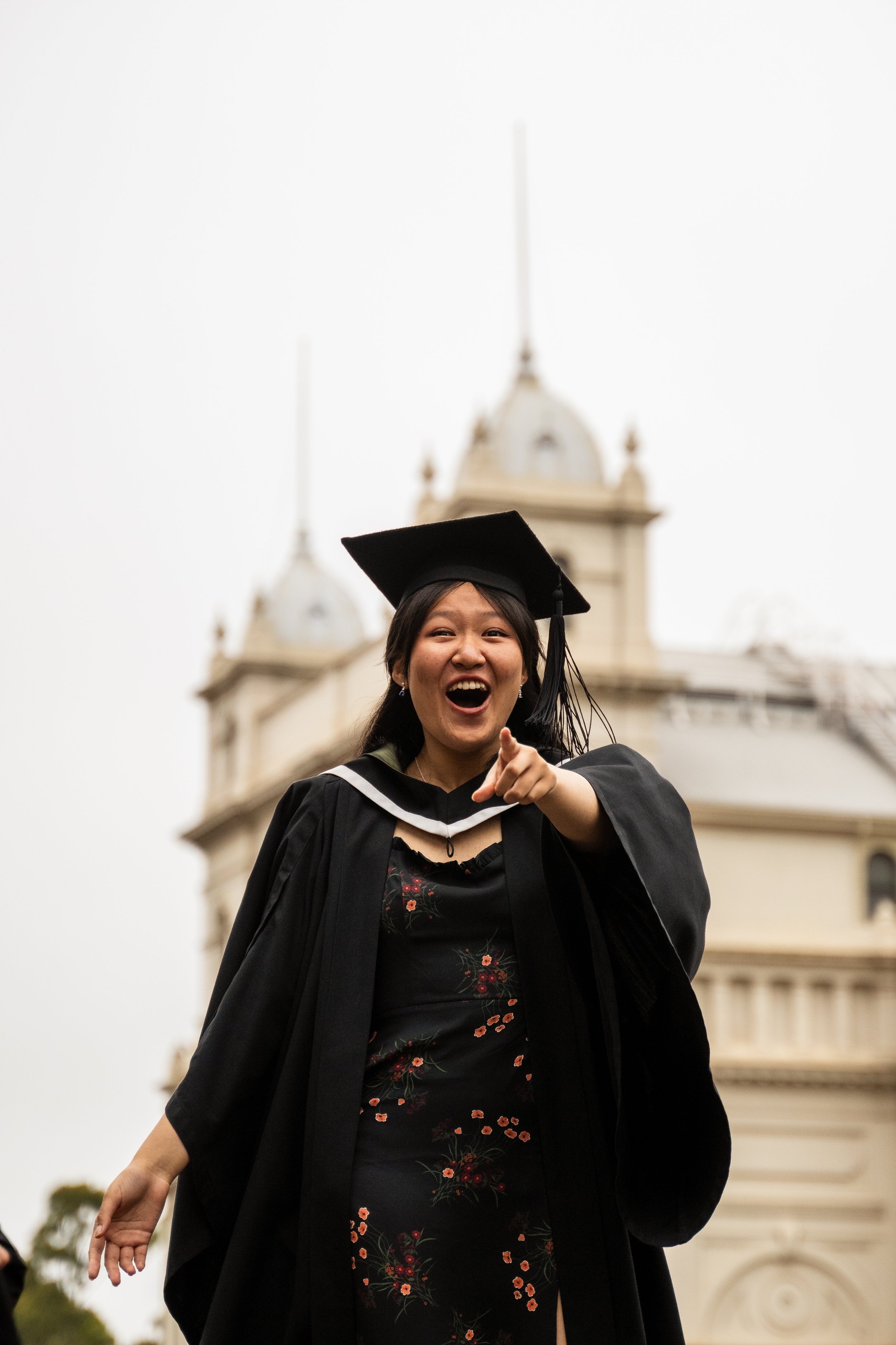 Happy young woman in graduation cap and gown pointing at the camera, standing outdoors with a historic building in the background.
