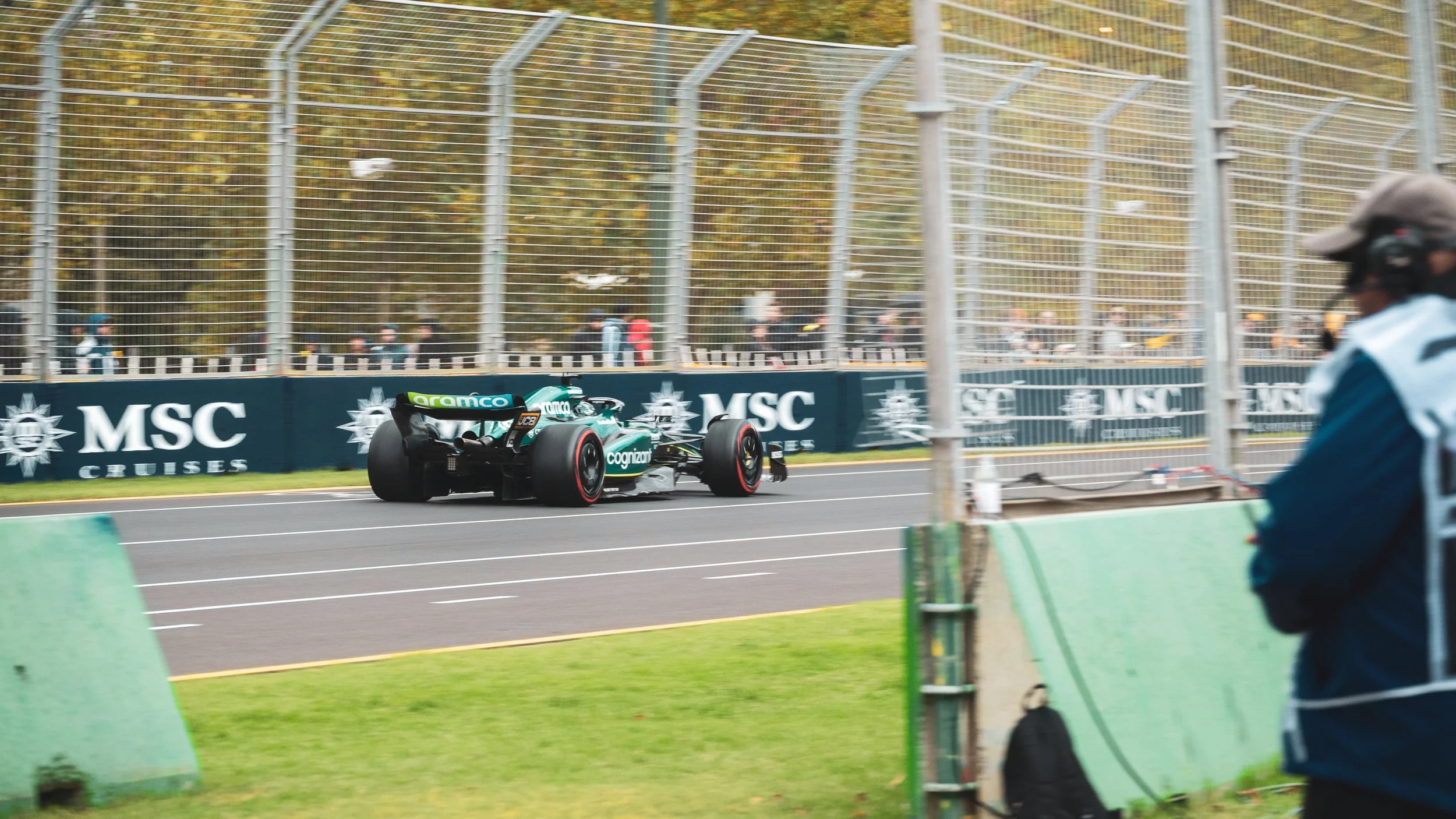 A racing car on a circuit track with a man watching, and barriers with MSI Cruises advertisement in the background.