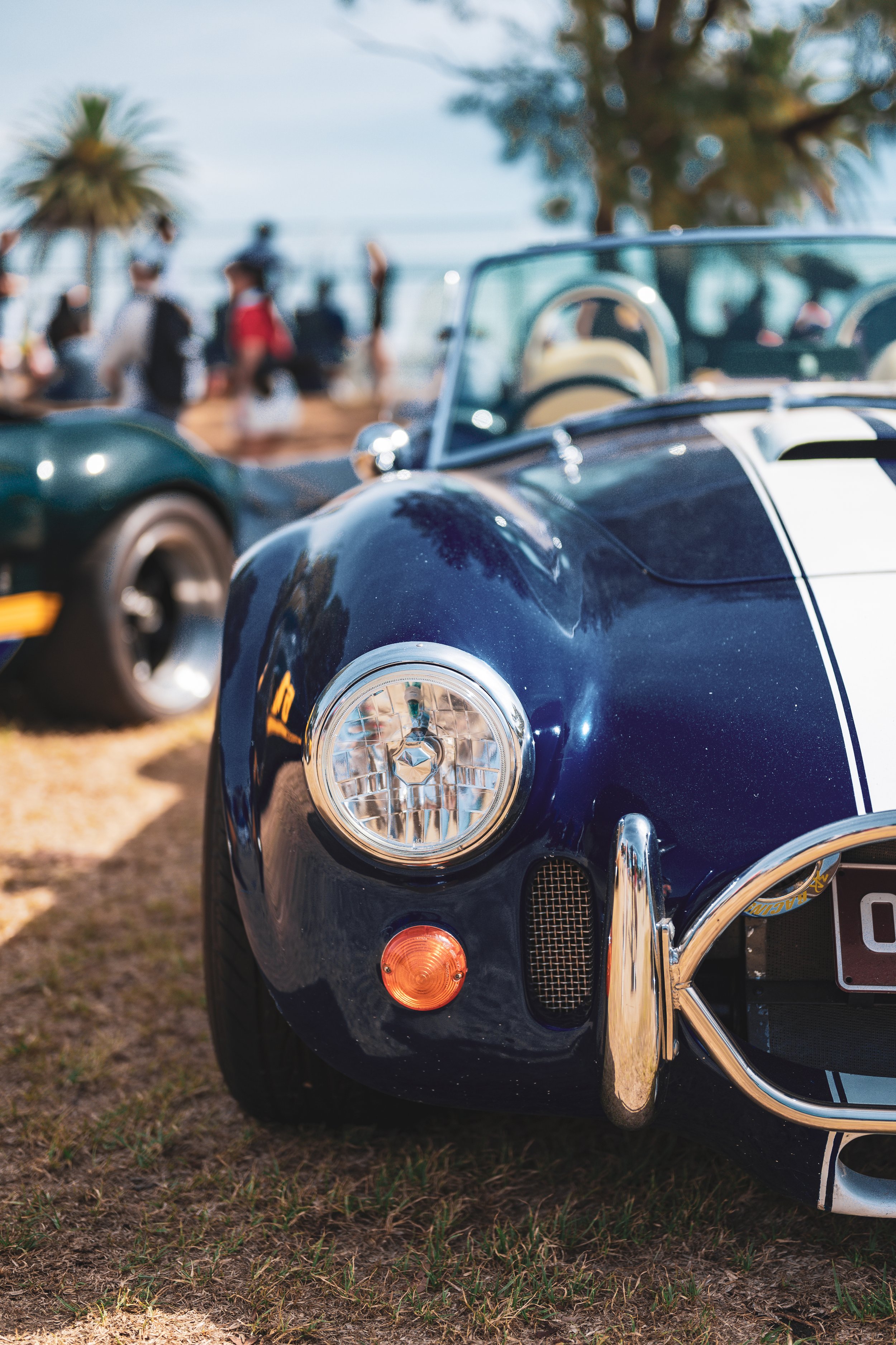 Close-up of a vintage dark blue convertible sports car with white racing stripes, parked outdoors at a car show. In the background, there are palm trees, a group of people, and another classic car, with a blue sky overhead.