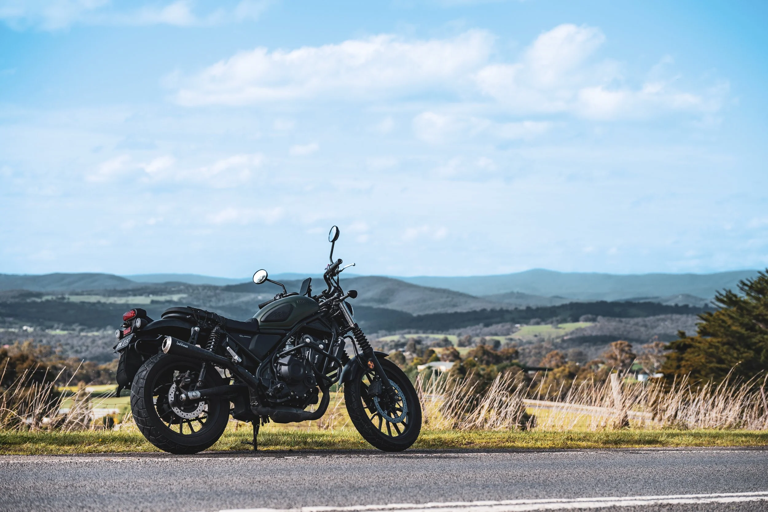 A black motorcycle parked on the side of a paved road with grassy fields and rolling hills in the background under a partly cloudy sky.