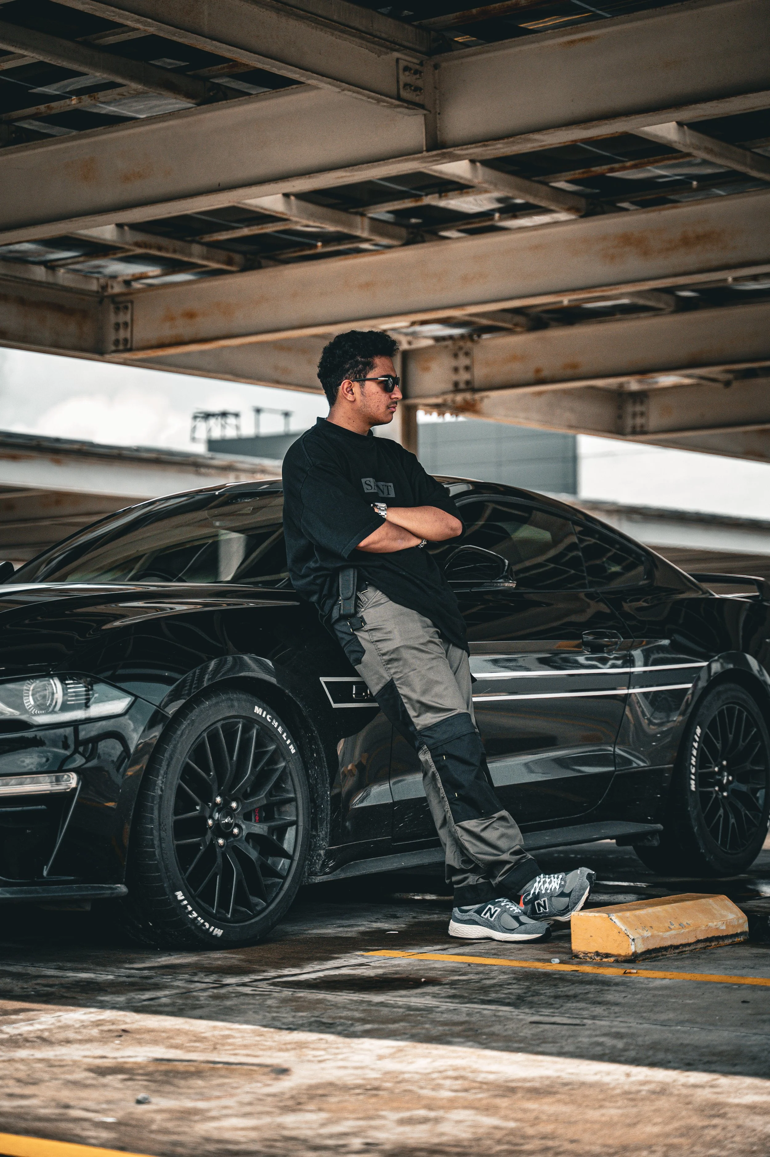 A young man wearing sunglasses, a black t-shirt, and gray cargo pants leaning against a black sports car in a parking lot under a metal structure.