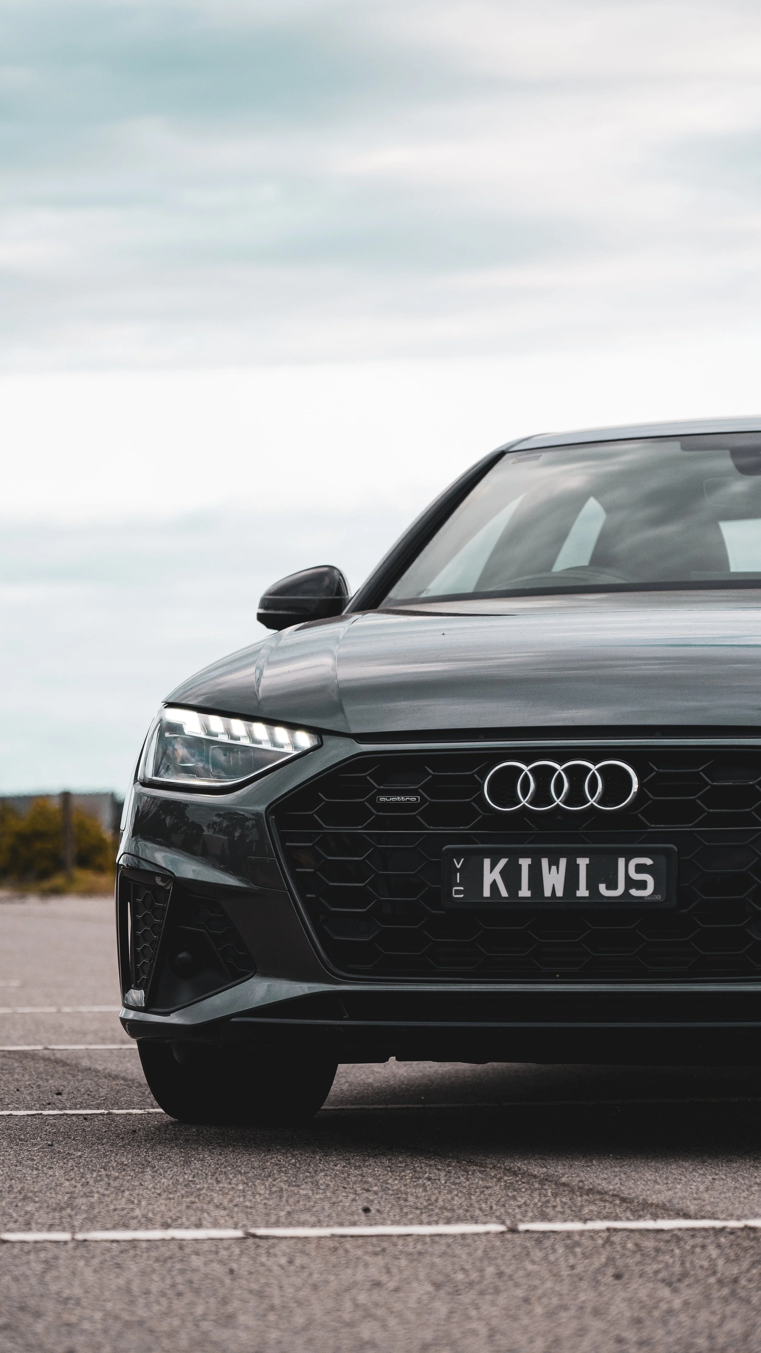 Front view of a black Audi car parked in an outdoor lot under cloudy sky, with a custom license plate reading "KIWJS".