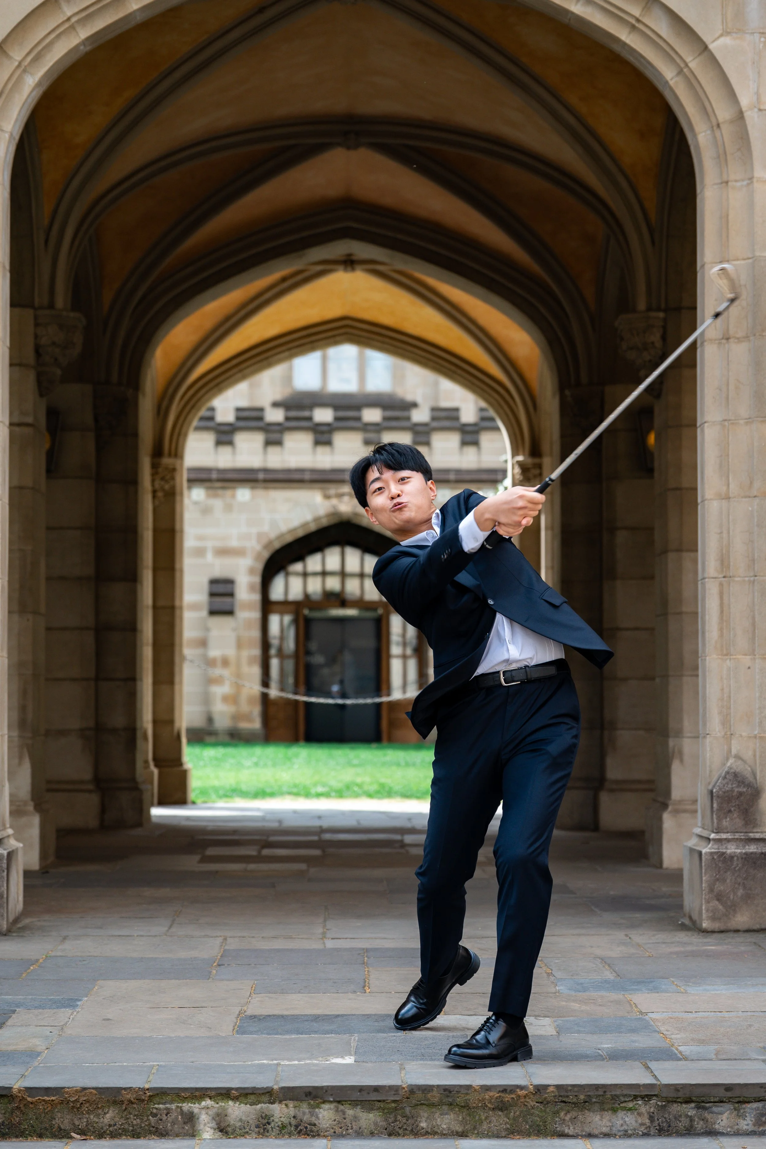 Young man in a business suit playing golf, swinging a golf club under a stone archway of a historic building.
