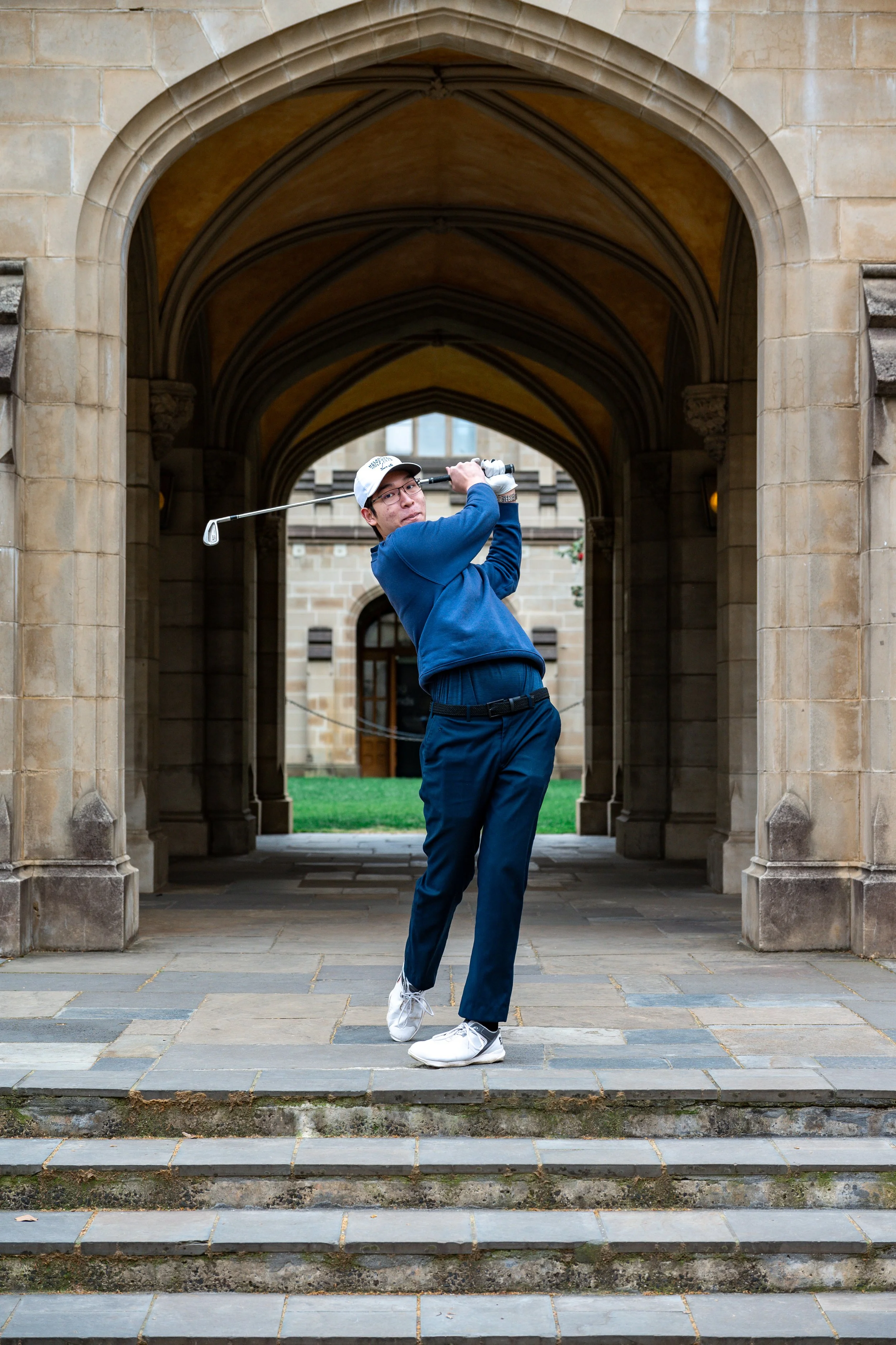 A man in athletic clothing and a baseball cap playing golf on stone steps under an ornate archway.