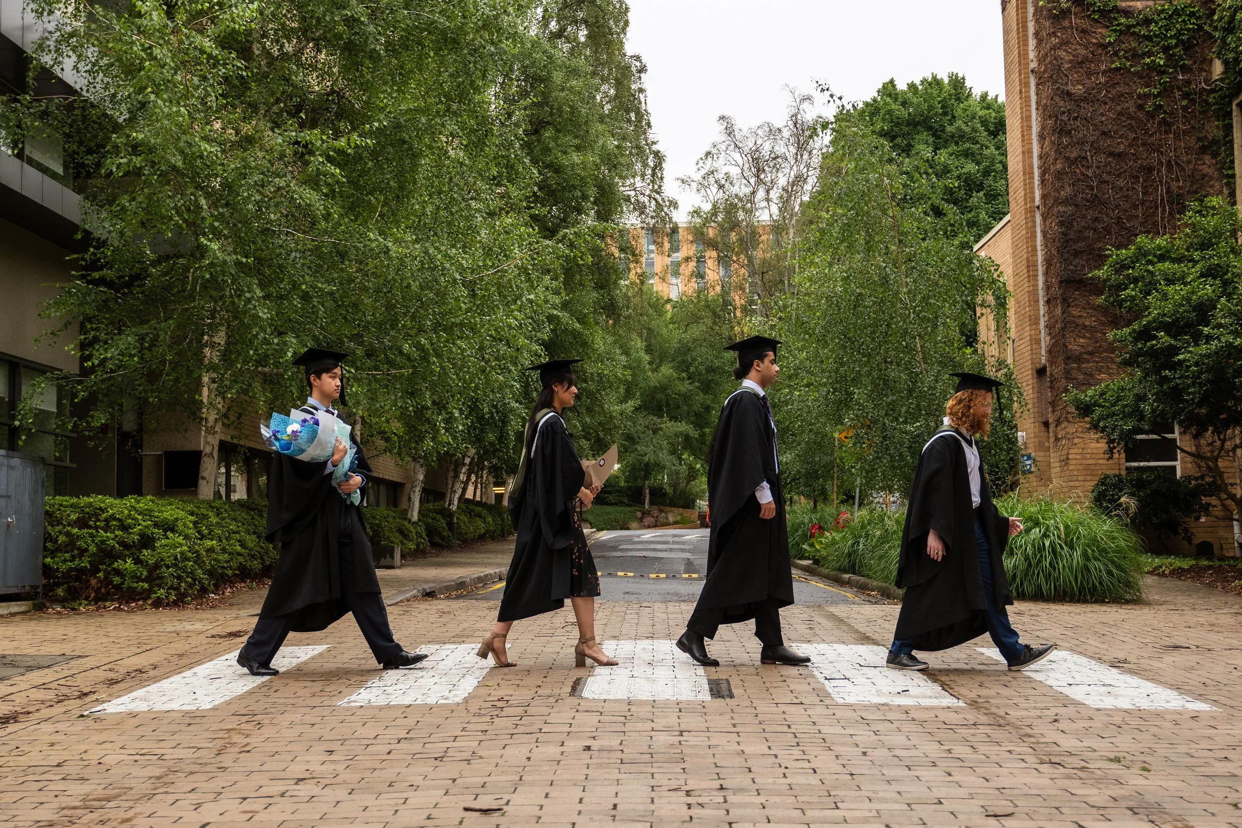 Group of five diverse graduates in black caps and gowns walking across a zebra crossing outdoors.