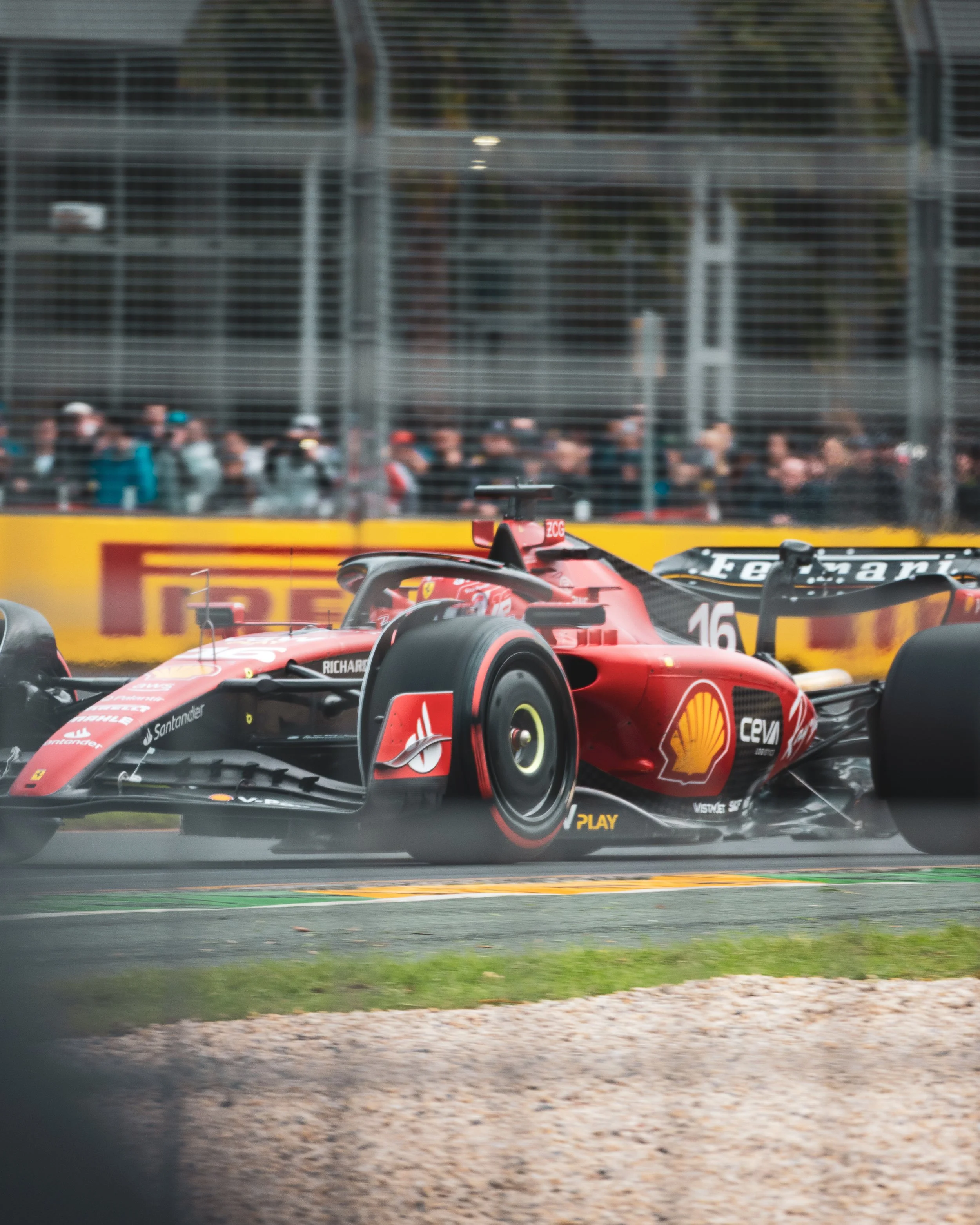 A red Formula 1 racing car on track during a race, with spectators and fencing in the background.