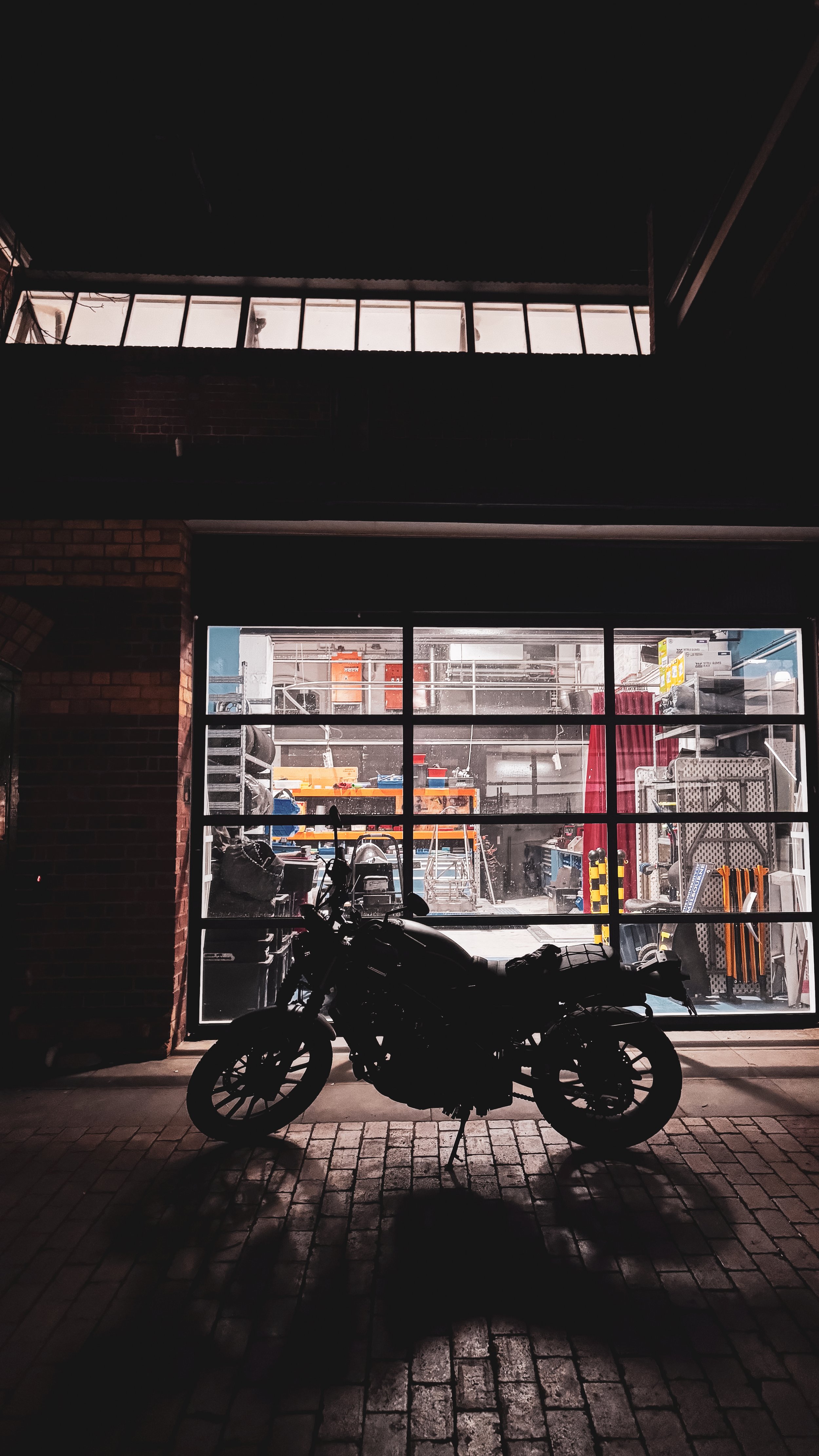 A black motorcycle parked in front of a garage with a large glass window, revealing a workshop with tools and equipment inside, lit from behind.