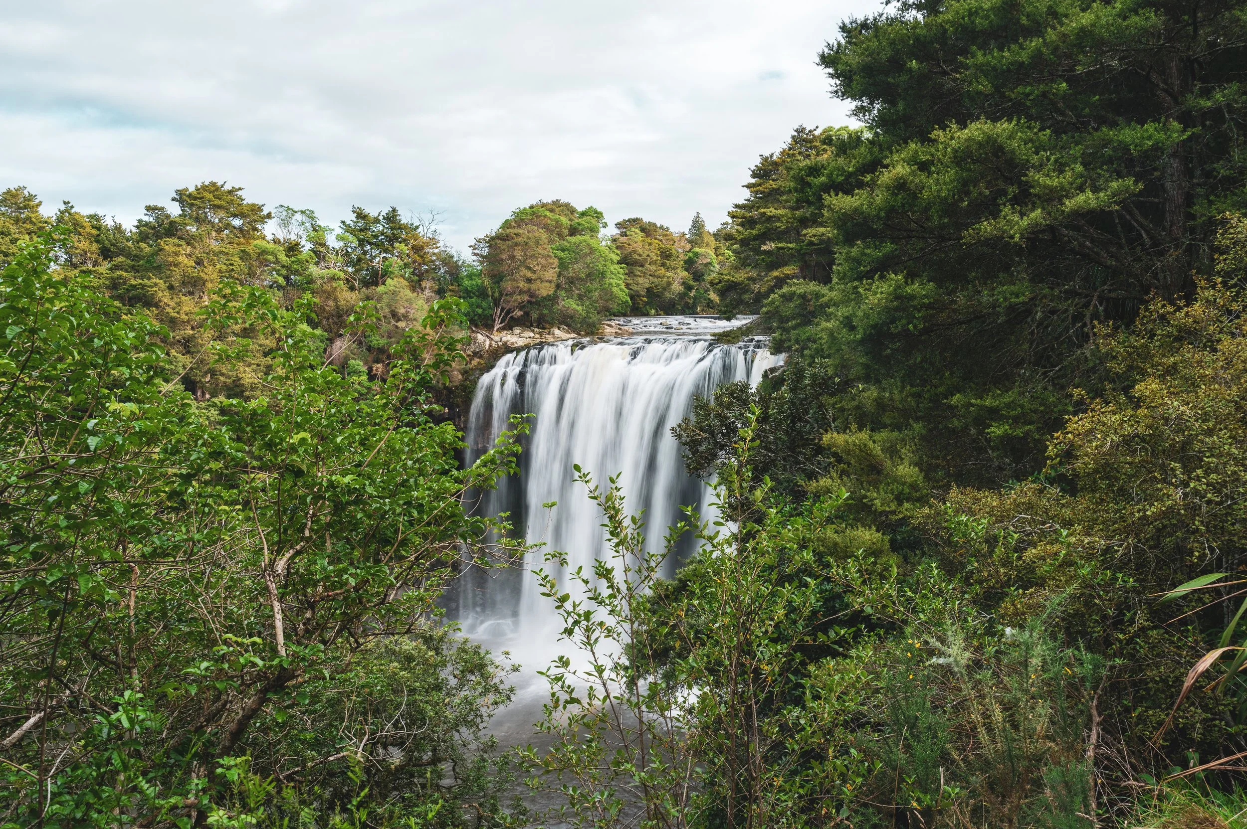 Waterfall flowing over rocks surrounded by lush green trees and bushes under cloudy sky