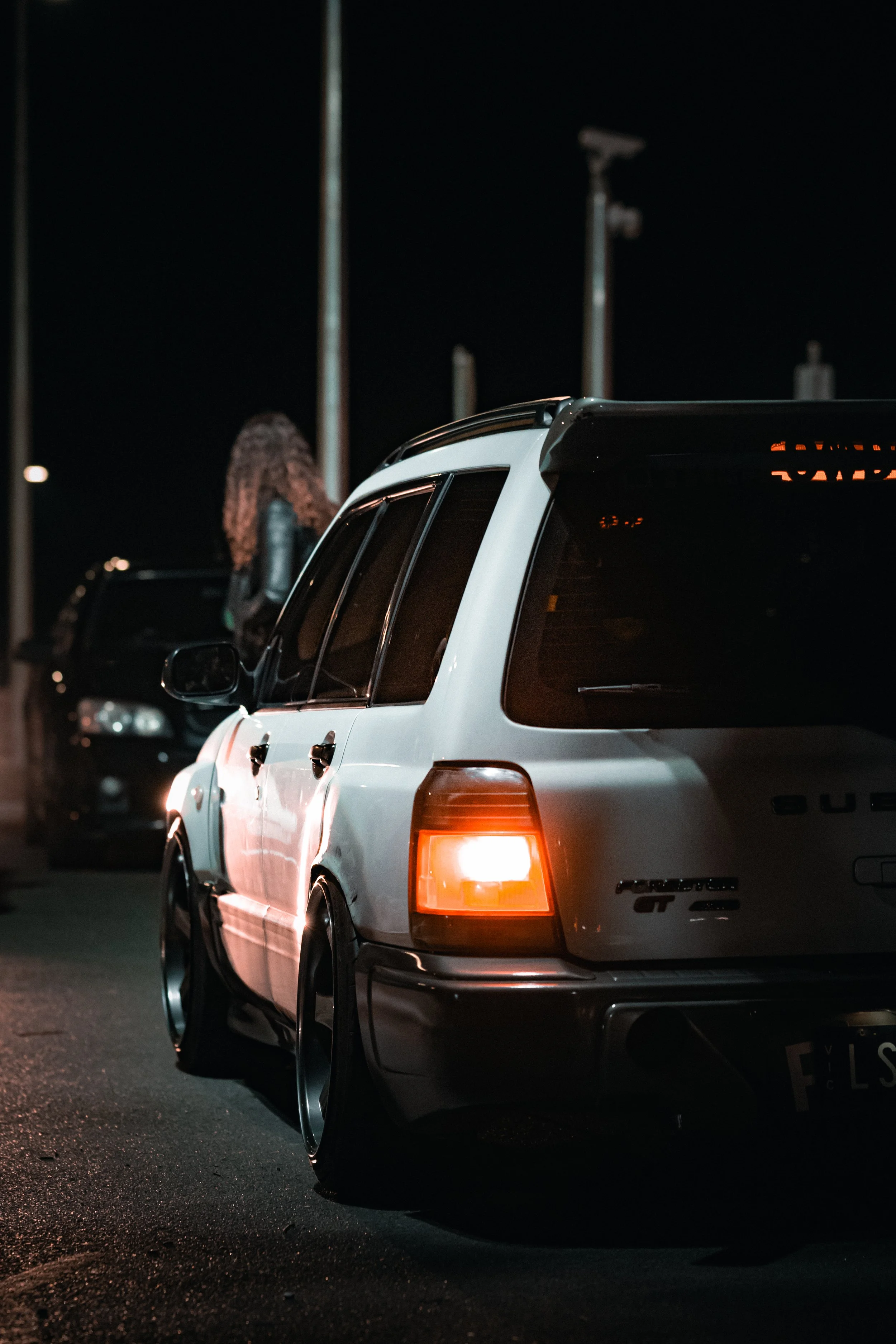 A white hatchback car with modified wheels parked at night, with a woman with curly hair standing beside it in the background.
