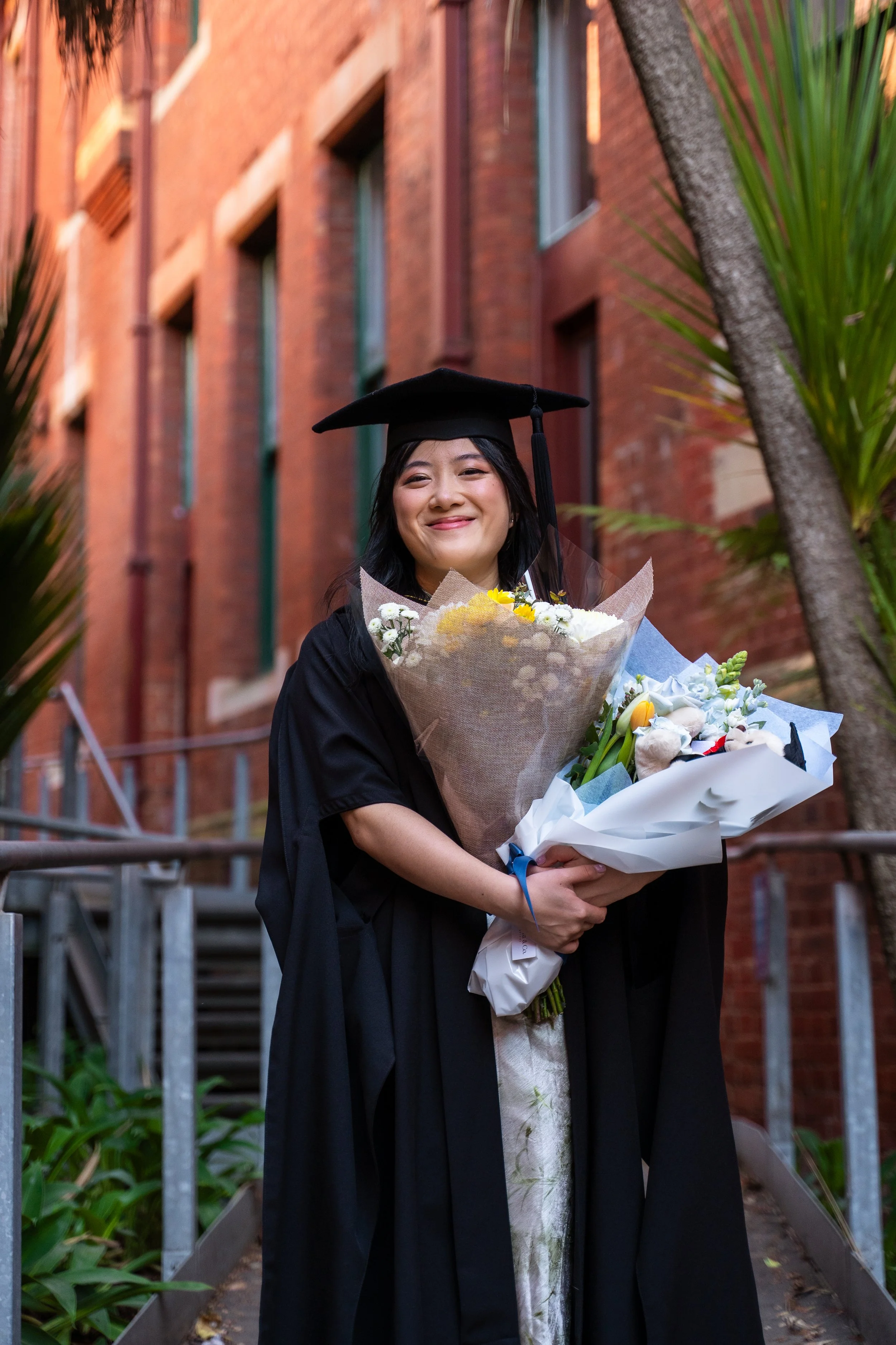 A young woman in a black graduation gown and cap holding a large bouquet of flowers, smiling, outdoors with a brick building and plants in the background.