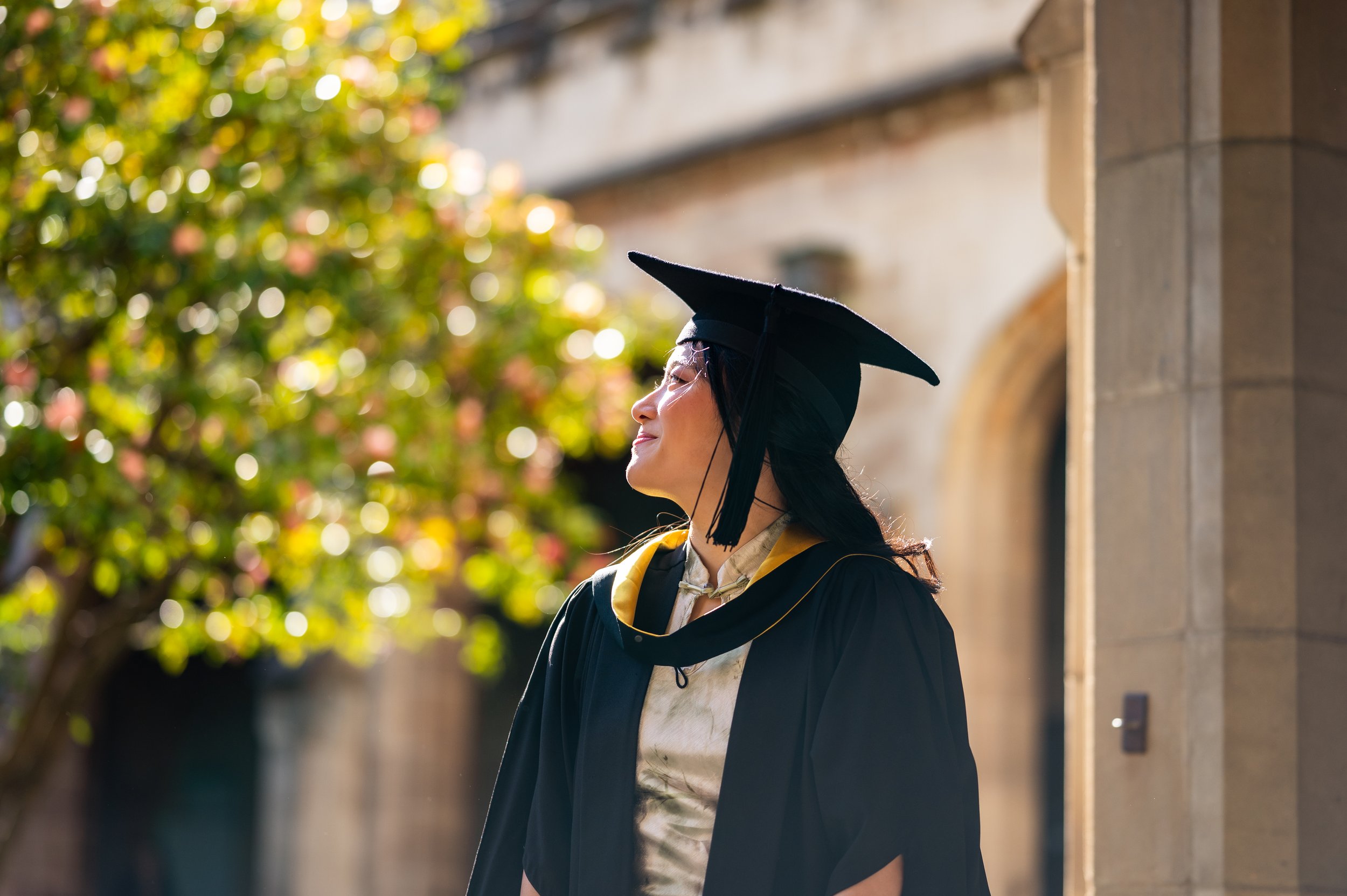 A woman in graduation cap and gown standing outdoors with sunlight and trees in the background.