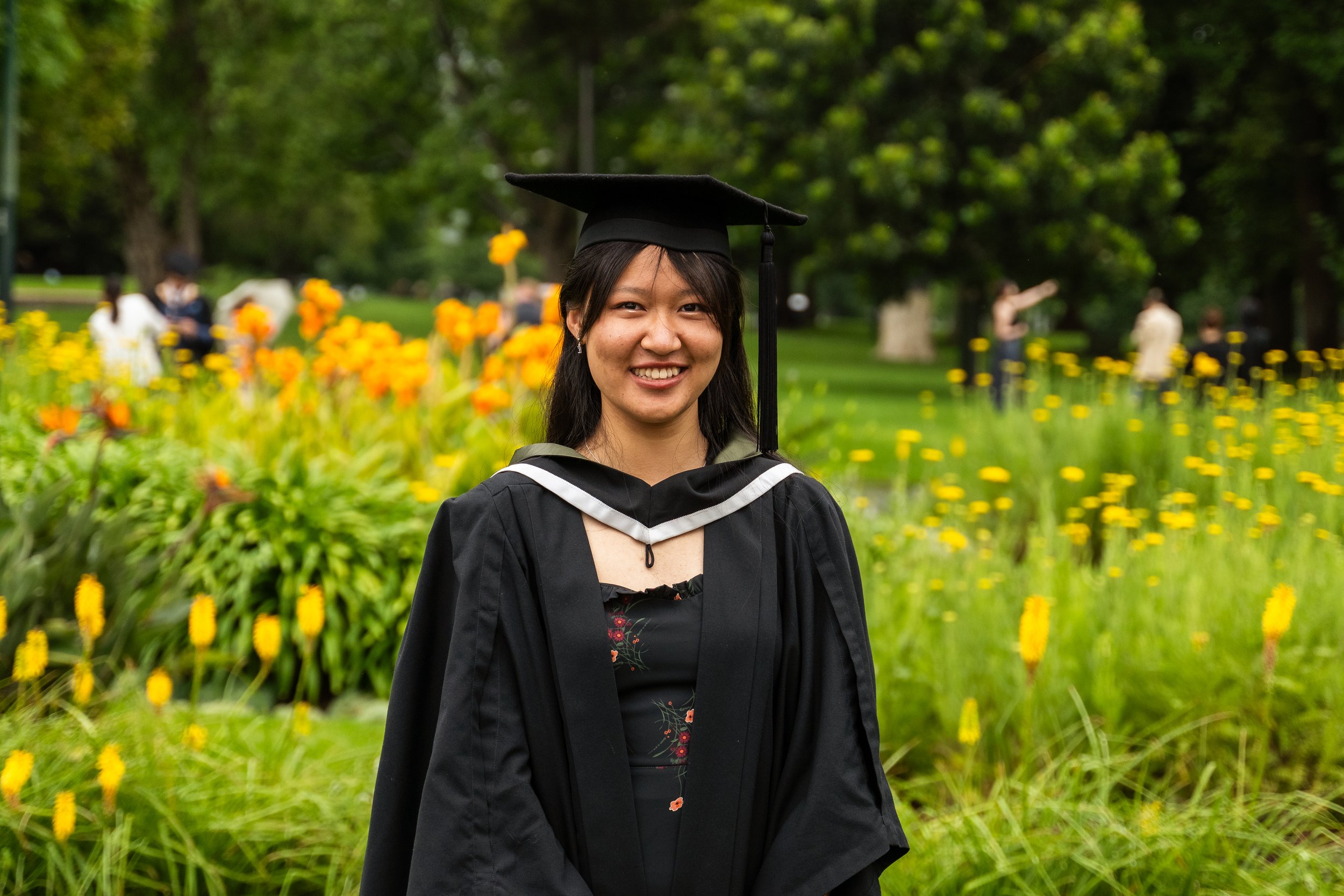 A young woman in a black graduation gown and cap standing outdoors on a lush green lawn with yellow flowers in the background, smiling at the camera.