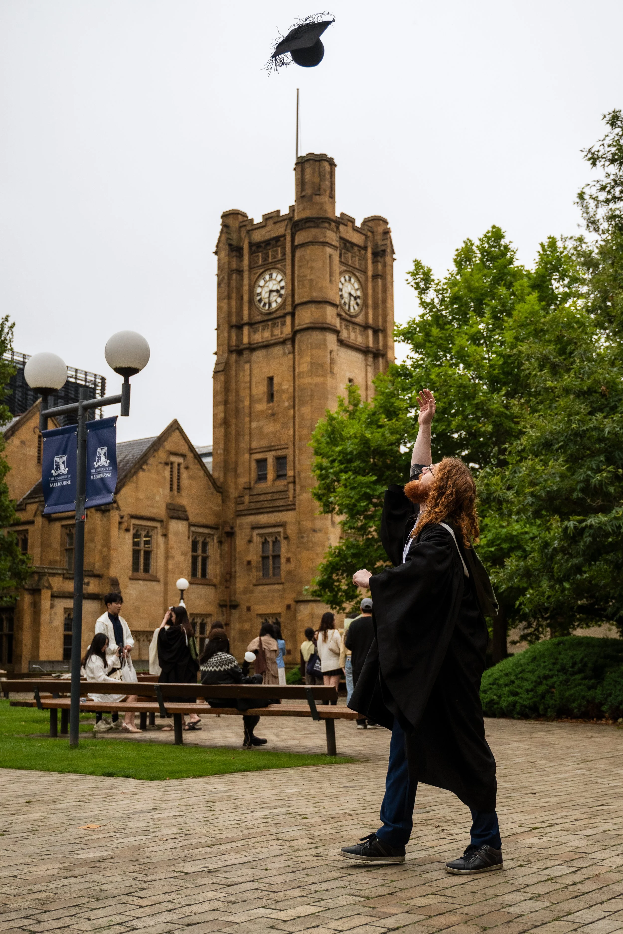 A graduate in a black cap and gown throws their cap into the air during a graduation celebration outside a historic university building with a clock tower.
