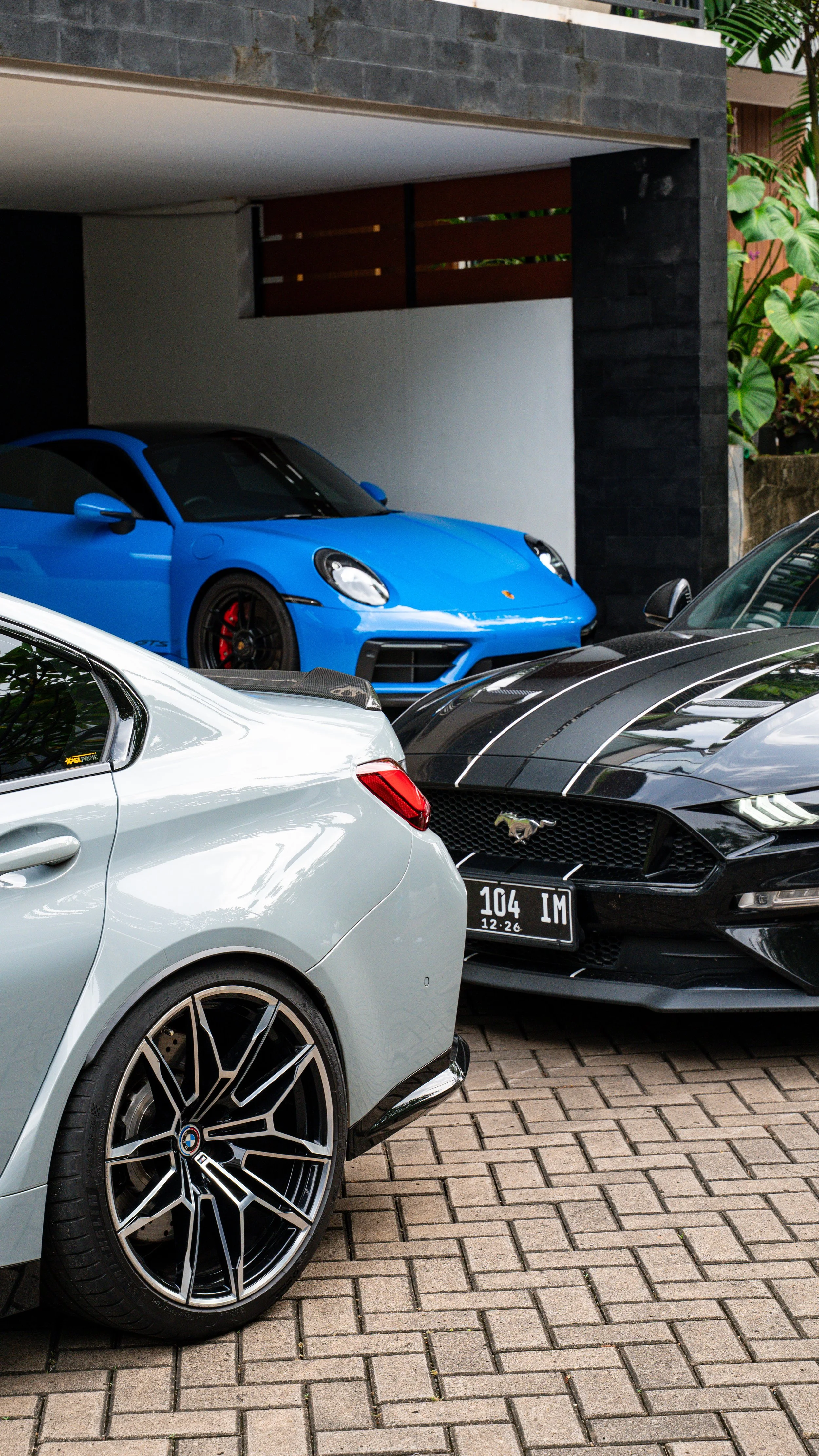 Three luxury cars parked in a shaded area, including a blue Porsche 911, a black Ford Mustang, and a silver BMW, with a modern building and greenery in the background.