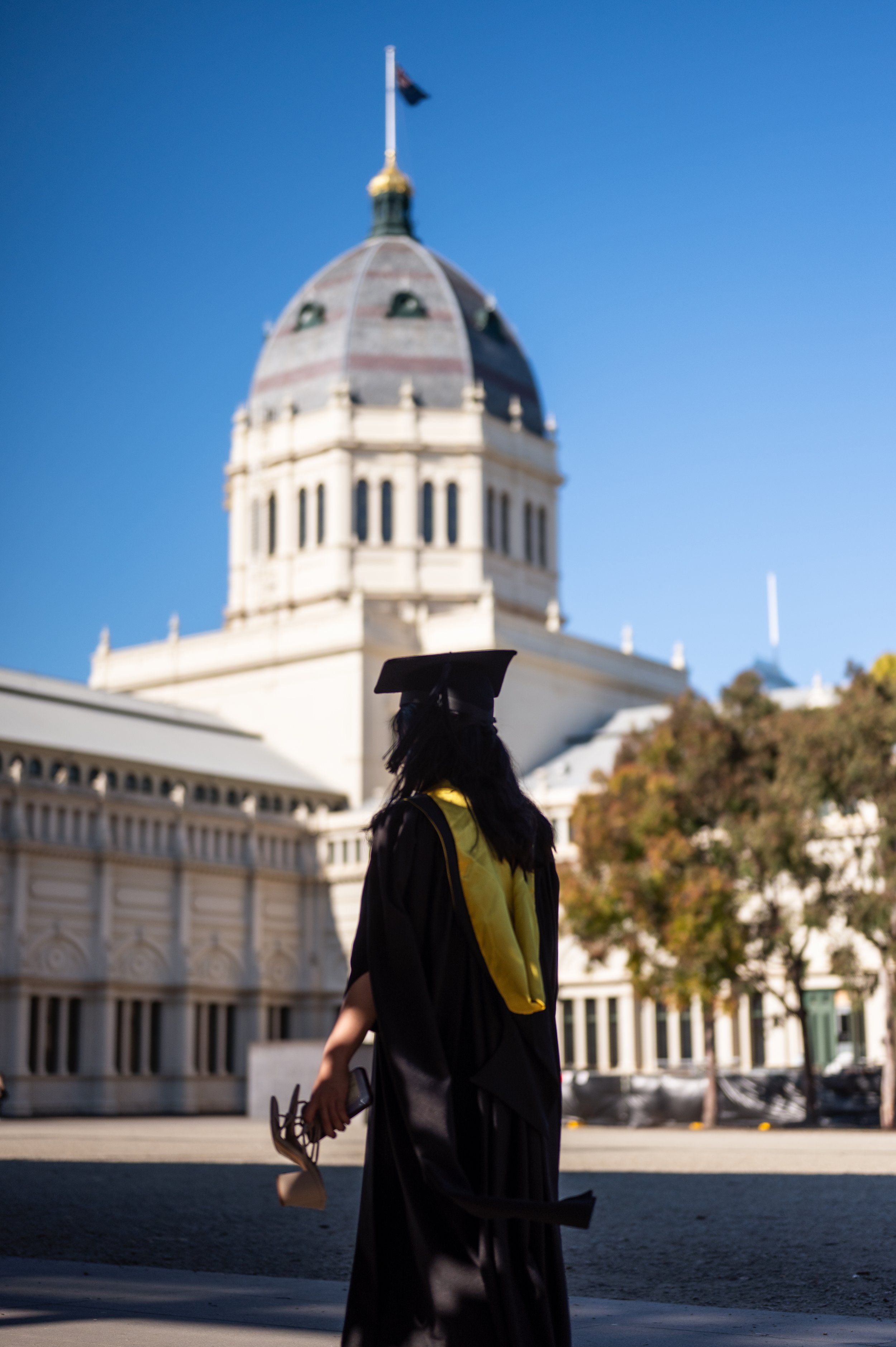 A person in a black graduation gown and cap with a yellow stole holding a diploma and a mortarboard, standing outdoors near a historic building with a domed roof and an American flag on top, against a bright blue sky.