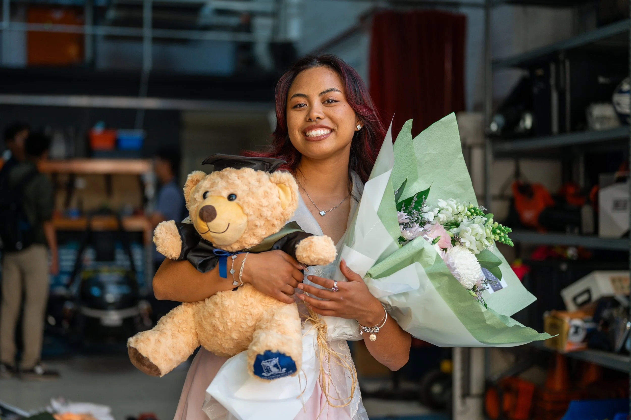 A young woman with dark red hair smiling, holding a large bouquet of flowers and a teddy bear with a graduation cap and gown in a garage or workshop setting.