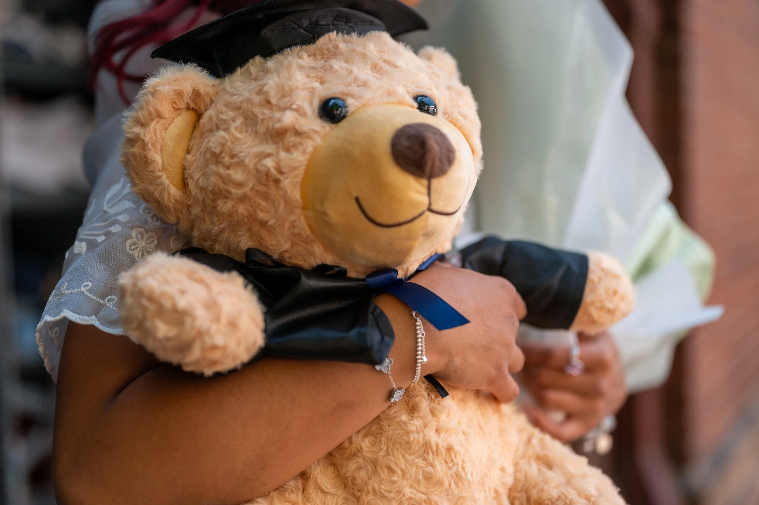 A plush teddy bear with light brown fur, wearing a black graduation cap and gown, being held by two people.