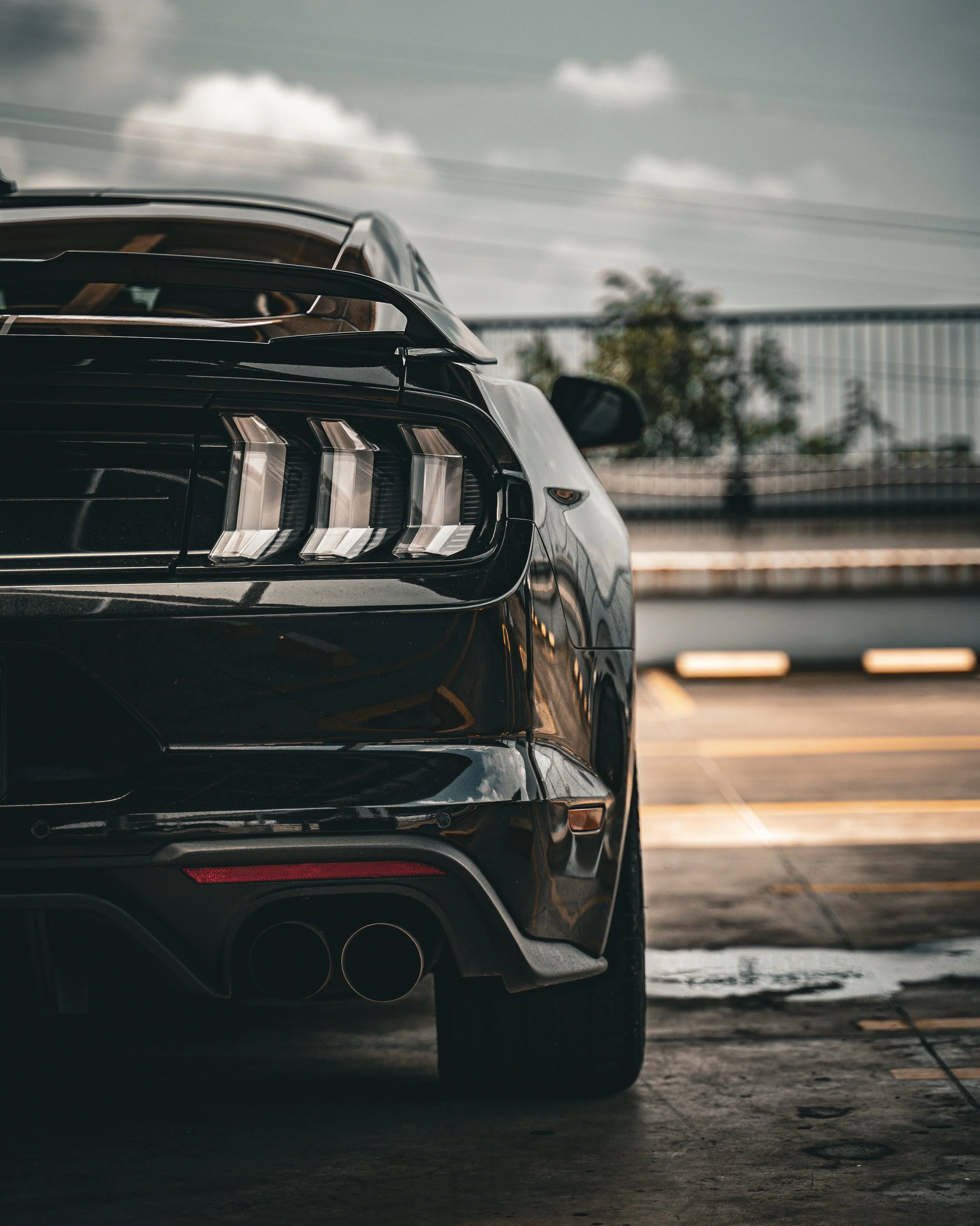 Close-up of the rear of a black Ford Mustang, showing taillights, dual exhaust pipes, and part of the rear wheel, parked in a lot with a cloudy sky in the background.