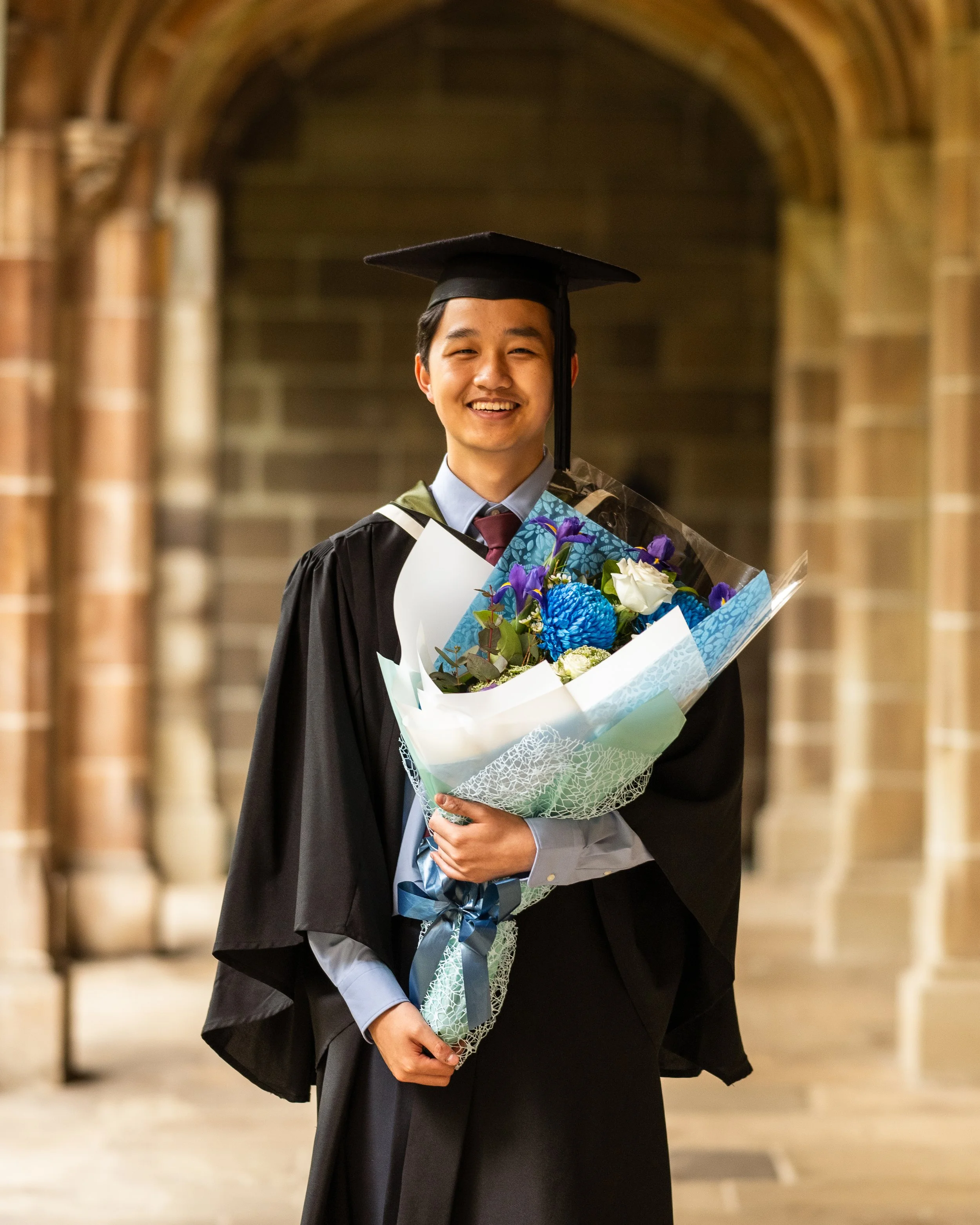 A young man in graduation cap and gown holding a bouquet of blue, purple, and white flowers inside a historic brick building.