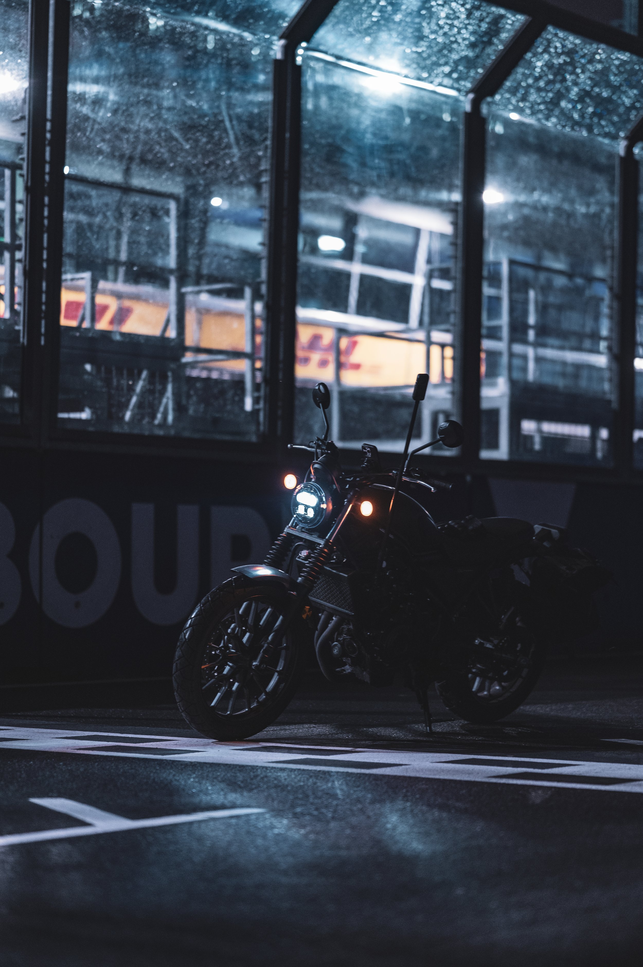A motorcycle parked in a dimly lit street at night with a bus stop shelter behind it, rain droplets on the shelter's glass, and wet pavement reflecting light.