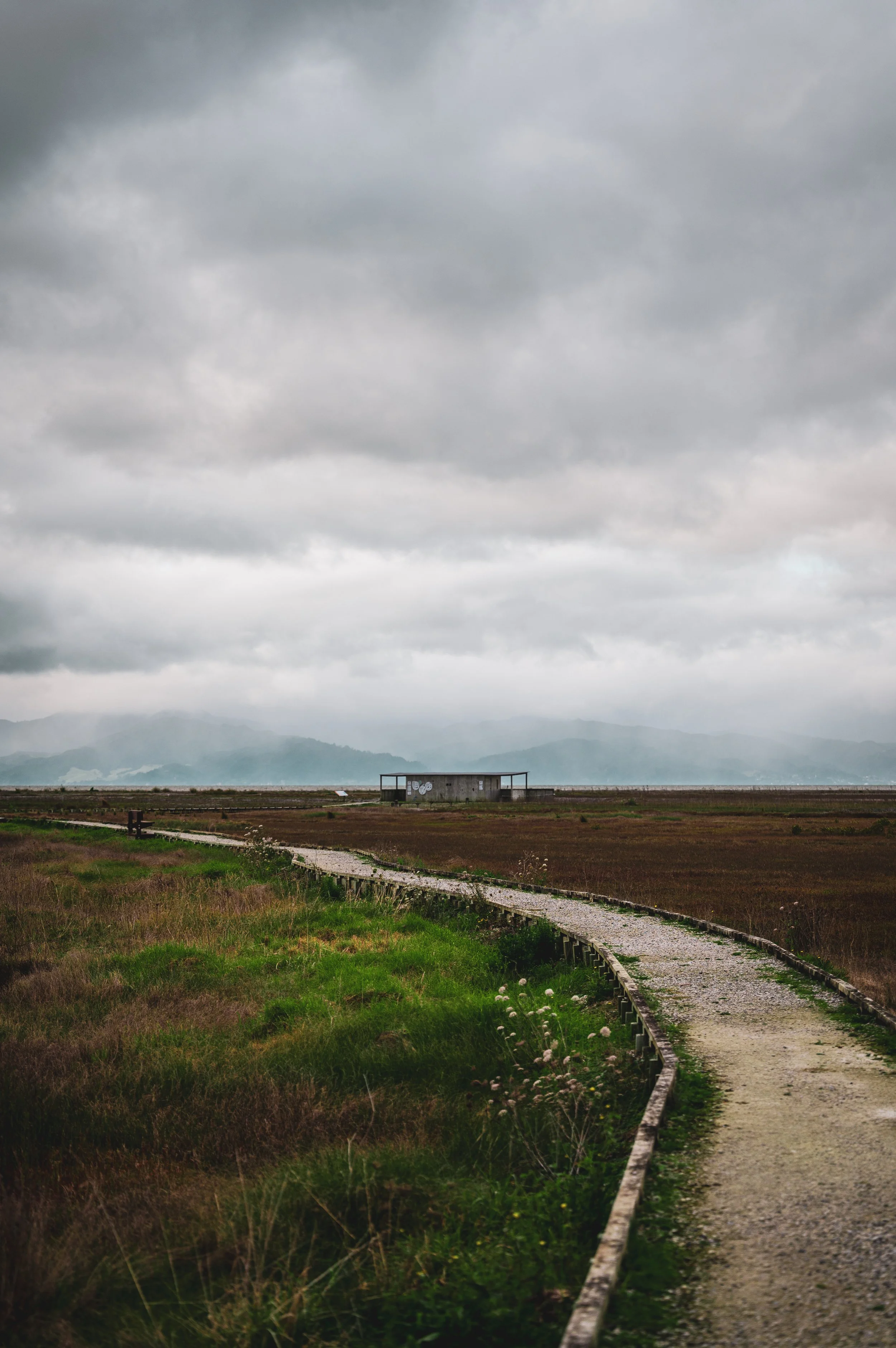A winding gravel path through a vast, flat landscape of grass and moor, with a small building in the distance under a cloudy, overcast sky.
