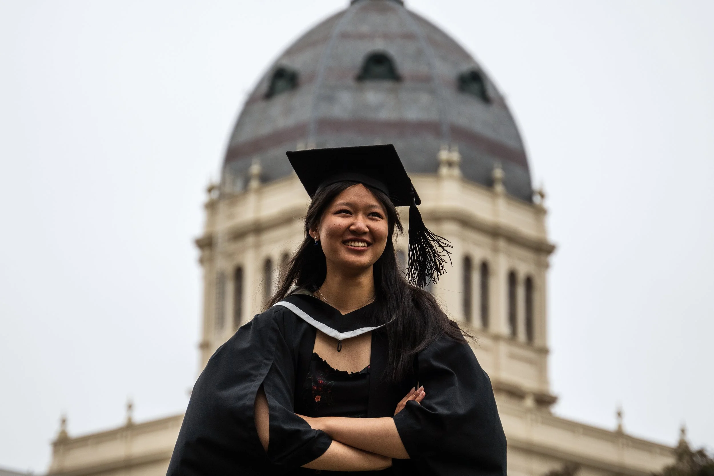 A young woman in a graduation gown and cap stands with arms crossed, smiling in front of a historic building with a large domed roof.