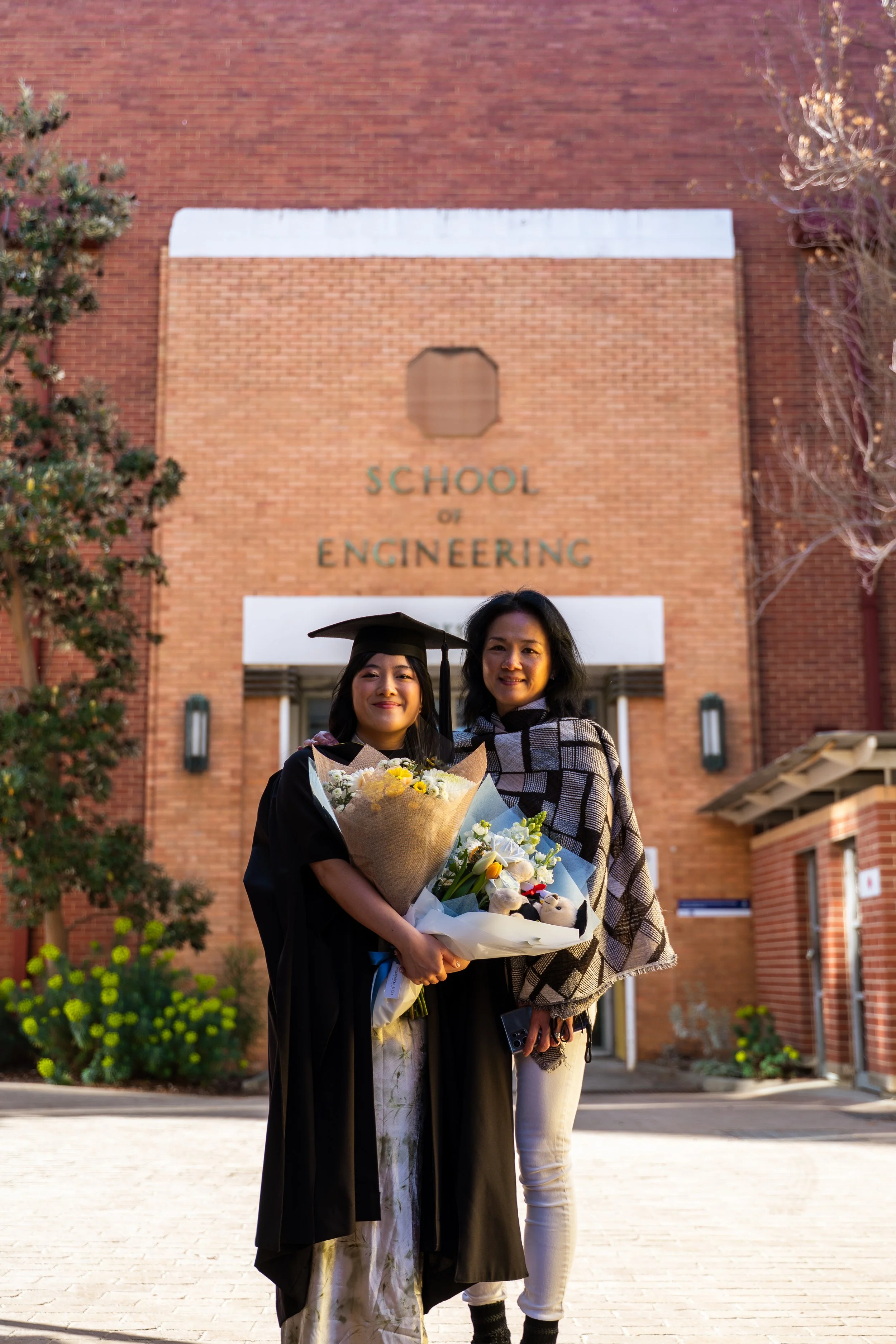 A woman in a graduation cap and gown holding a bouquet of flowers, standing next to another woman holding flowers, in front of a brick building with 'School of Engineering' sign.