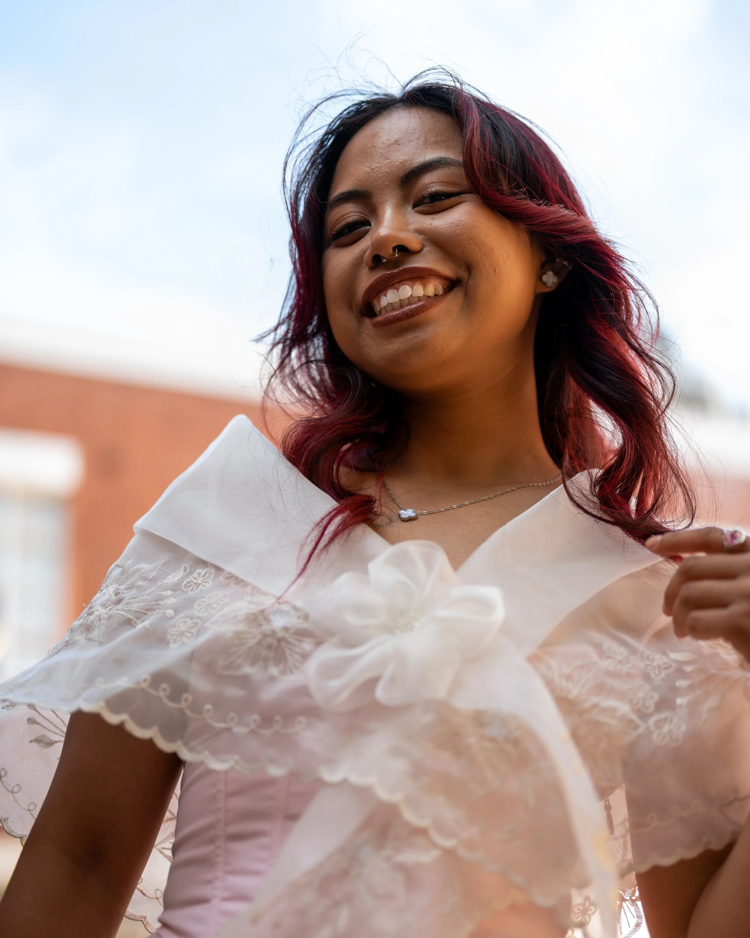 A young woman with shoulder-length hair dyed red at the ends, smiling outdoors against a blue sky and brick building, wearing a white lace dress with floral details and a necklace.