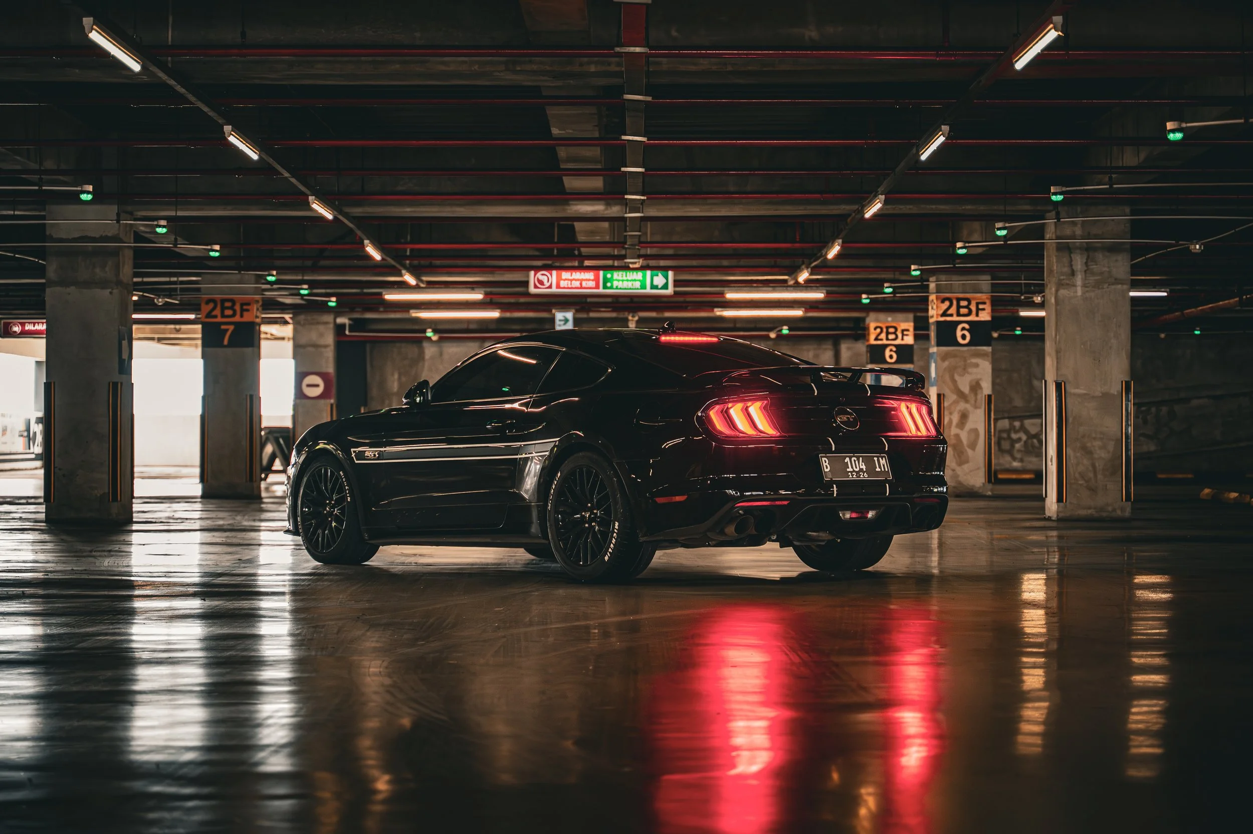 A black Ford Mustang parked in an underground parking garage with dim lighting, concrete pillars, and neon signs overhead.