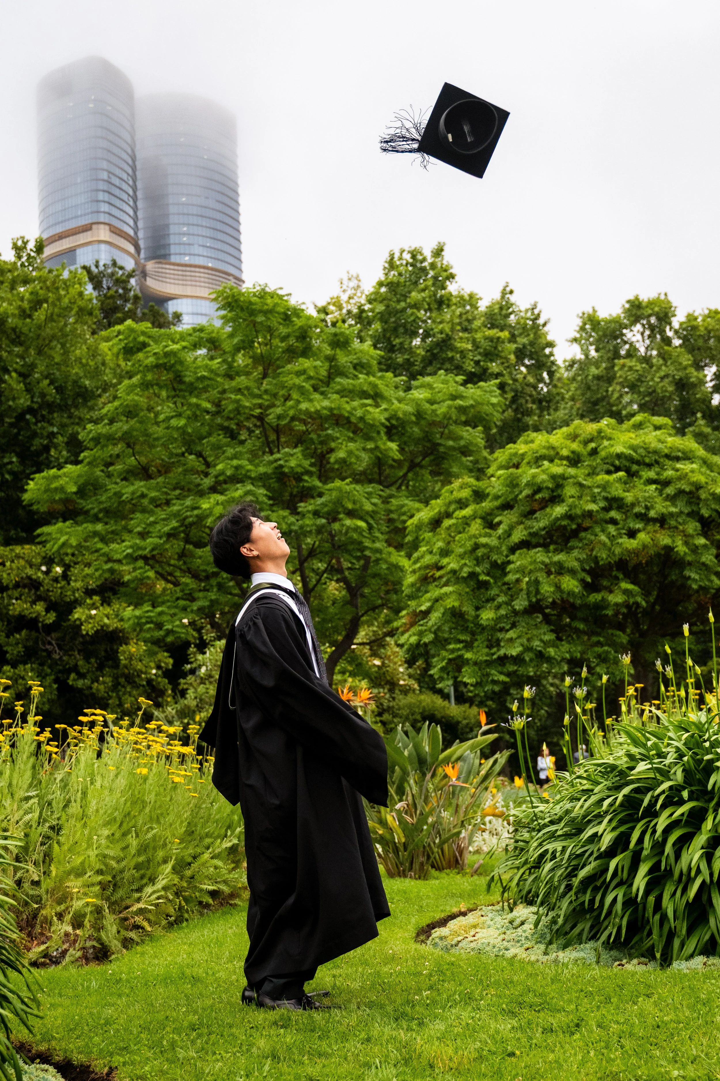 A young man in a graduation gown and cap throws his cap into the air in a park with green trees, yellow flowers, and tall buildings in the background.