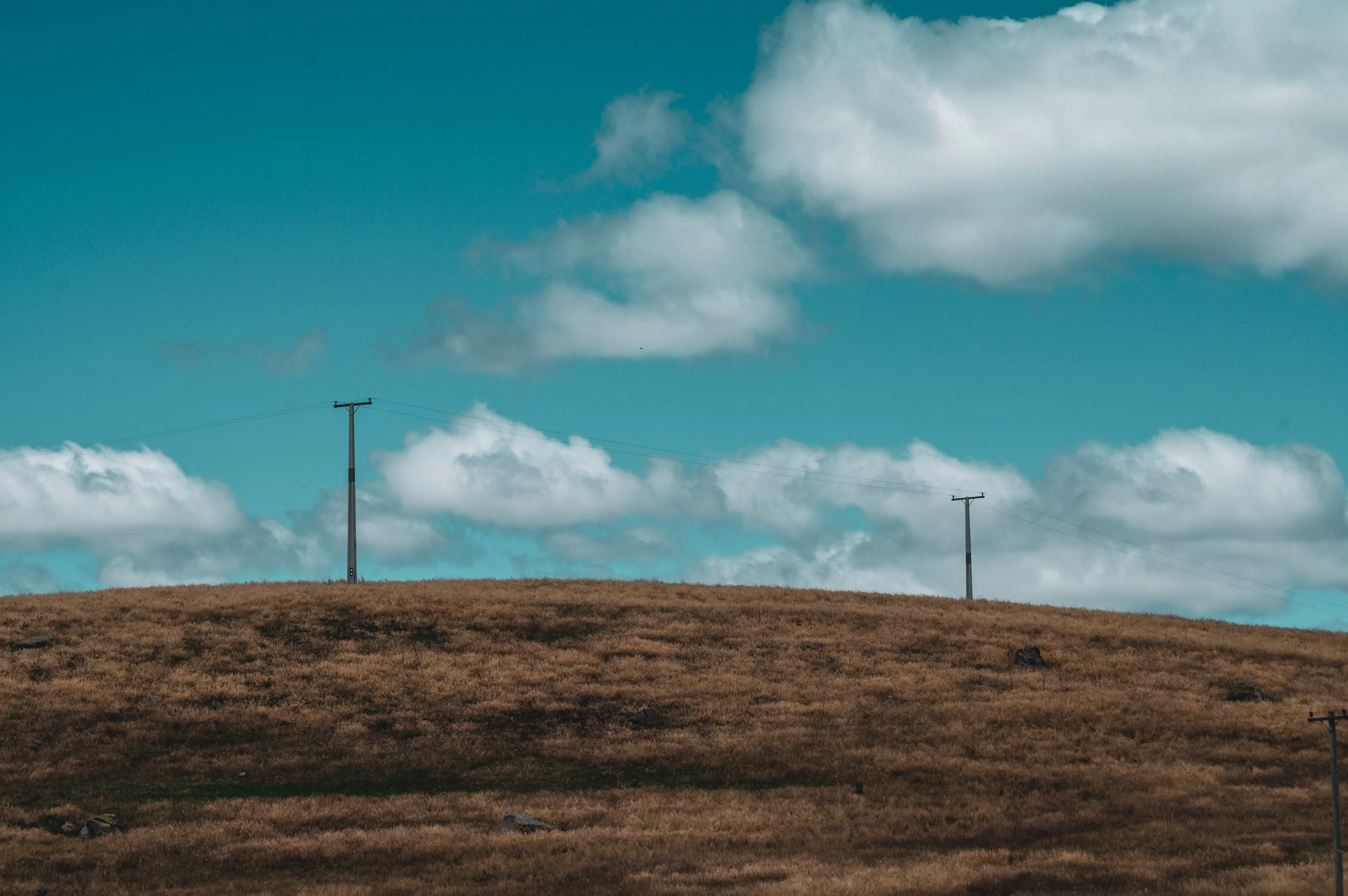 A rural landscape with a grassy hill and three utility poles under a sky with scattered clouds.