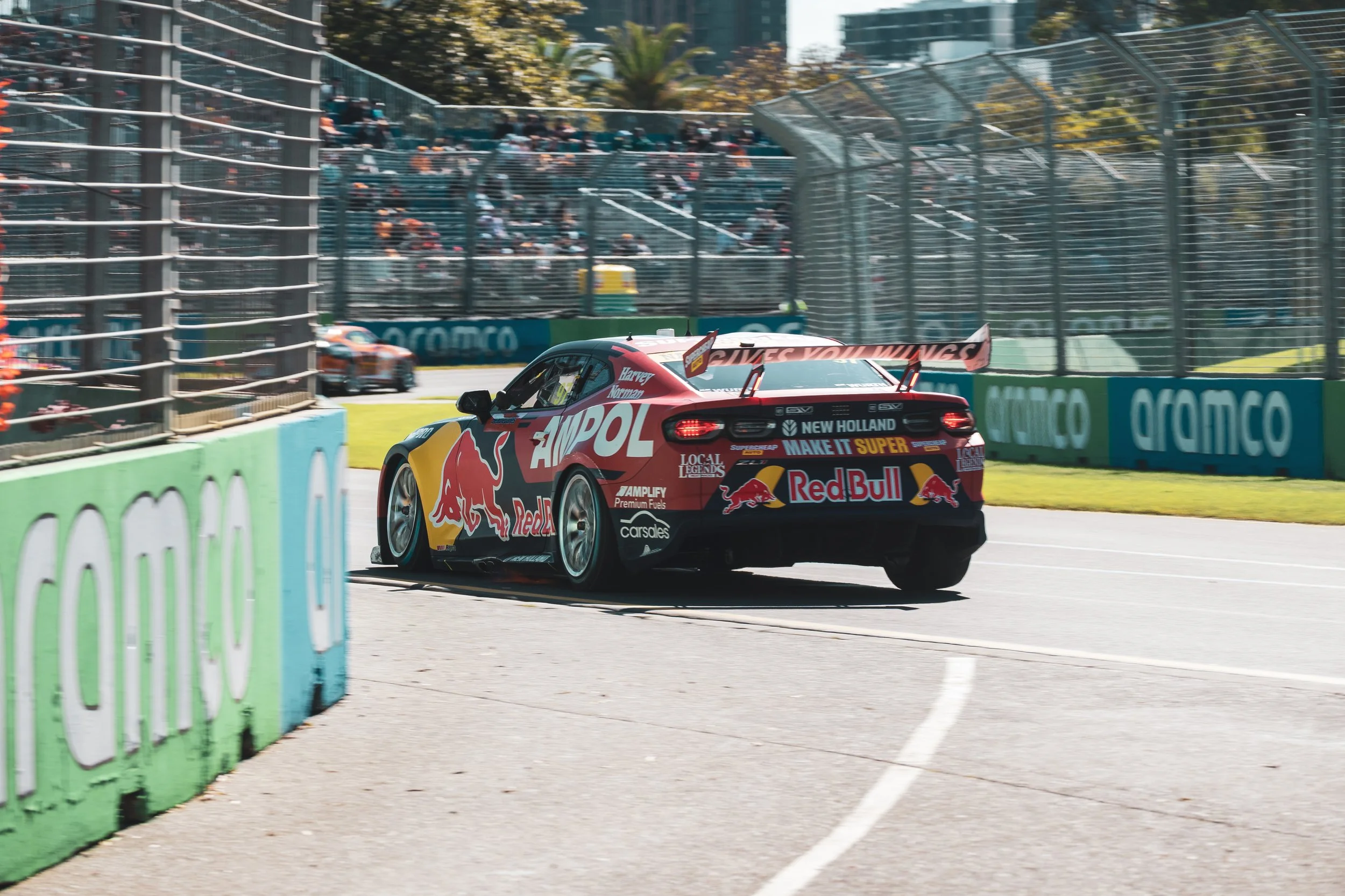 A race car on a race track with spectators in the stands and a green barrier with sponsor logos.