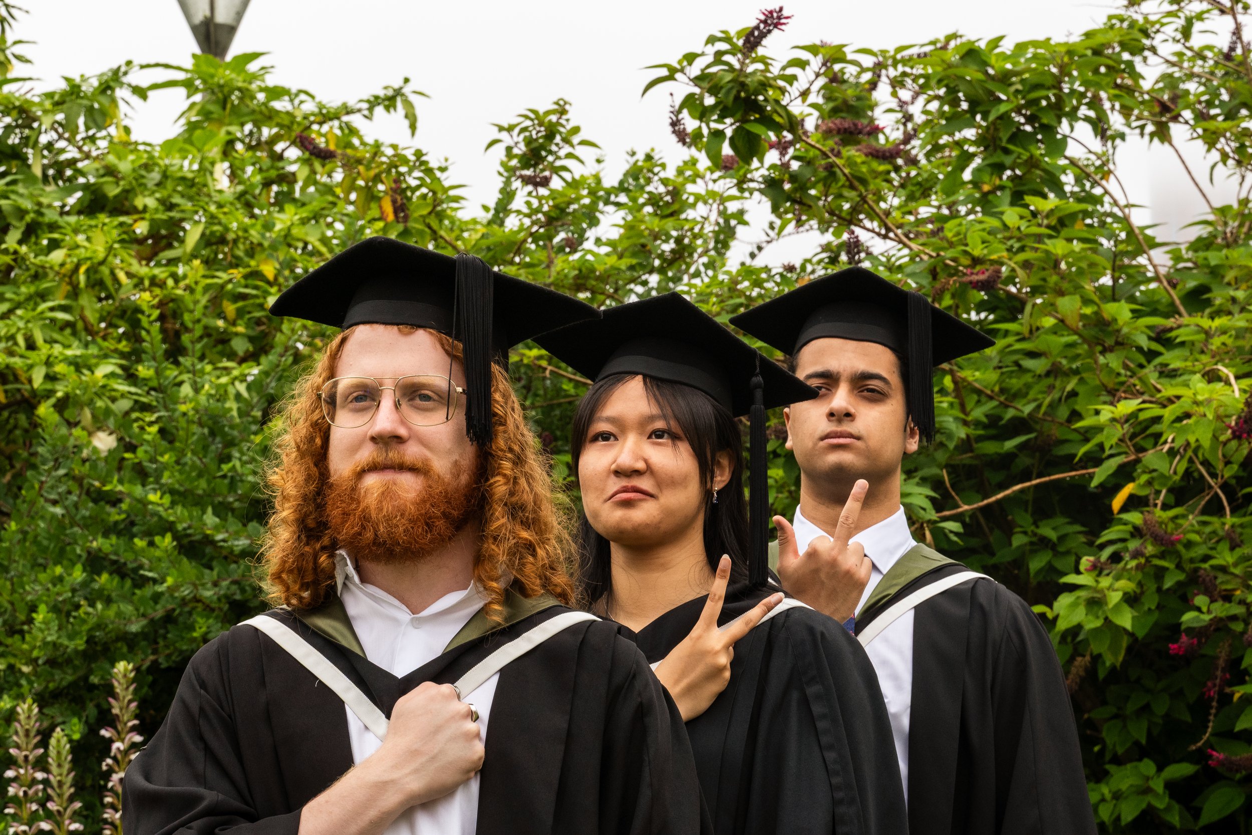 Three diverse graduates wearing caps and gowns making silly and serious facial expressions outdoors in front of green bushes.