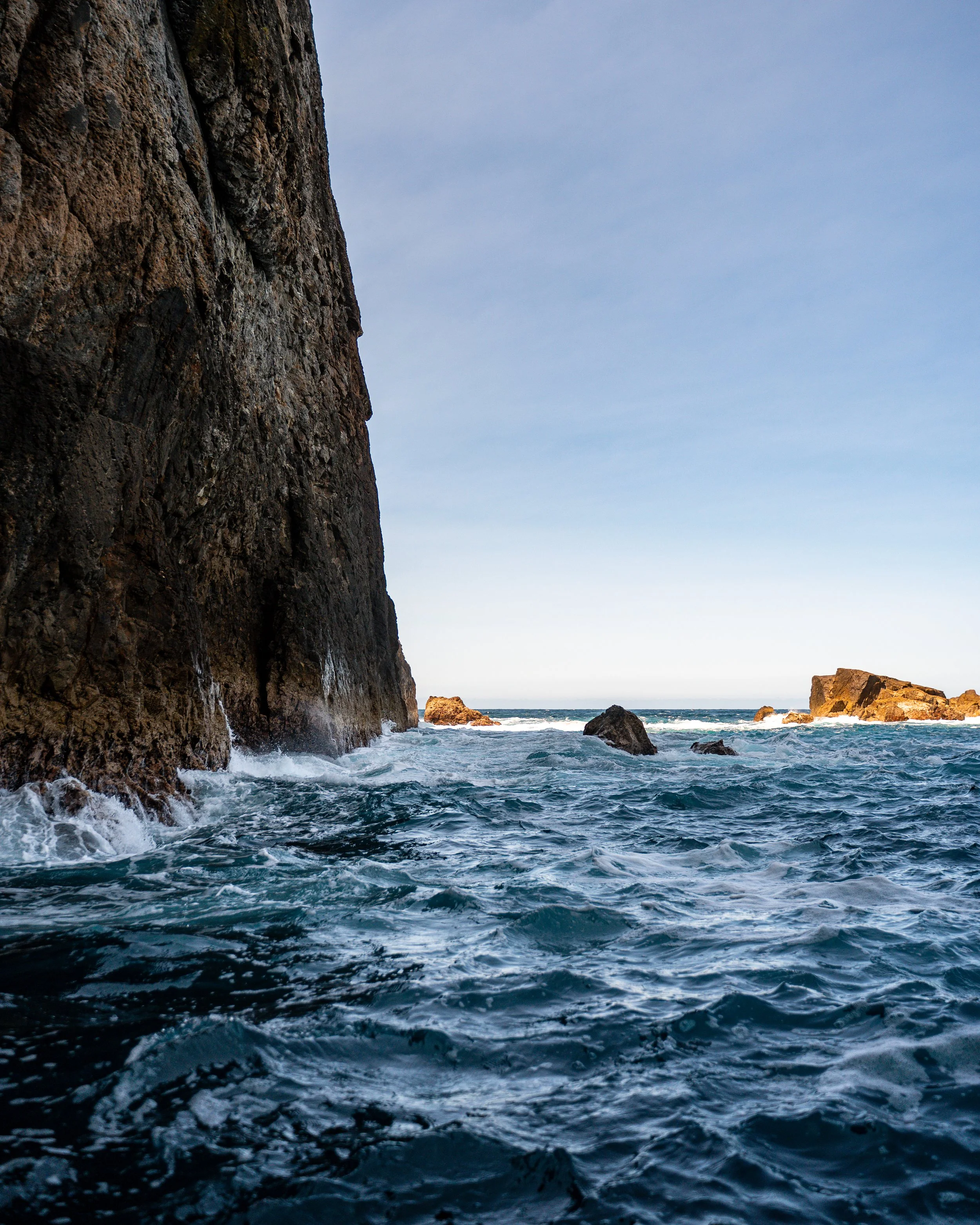 Cliffs on a rocky coastline with the ocean at the base and several rocks in the water, under a clear sky.
