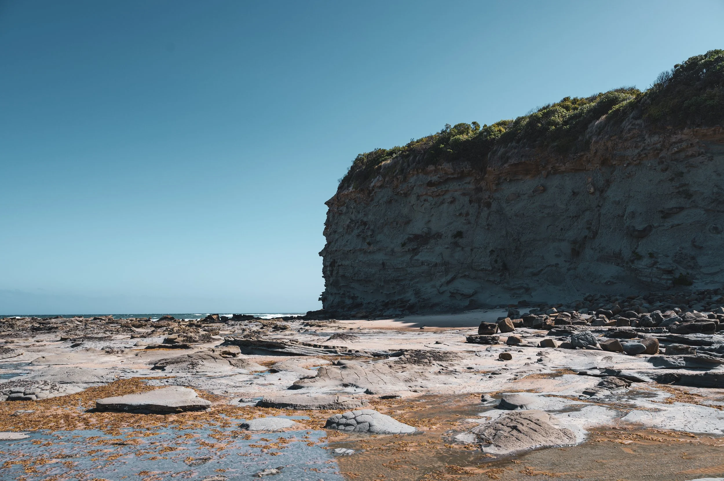 A rocky beach with a large cliff and green foliage on top, under a clear blue sky.
