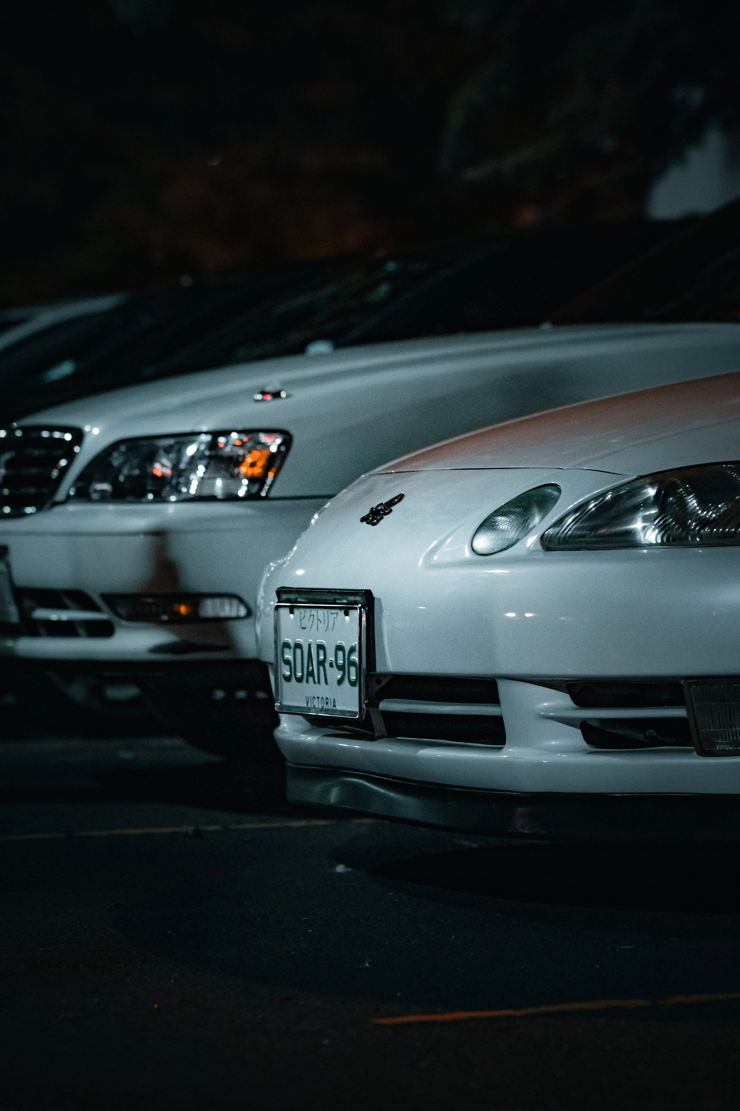 A lineup of three cars parked in a dark parking lot at night. The front car is a white sports car with a visible license plate reading 'SOAR-96' and a small sticker on the hood. The second car behind it is a white sedan.