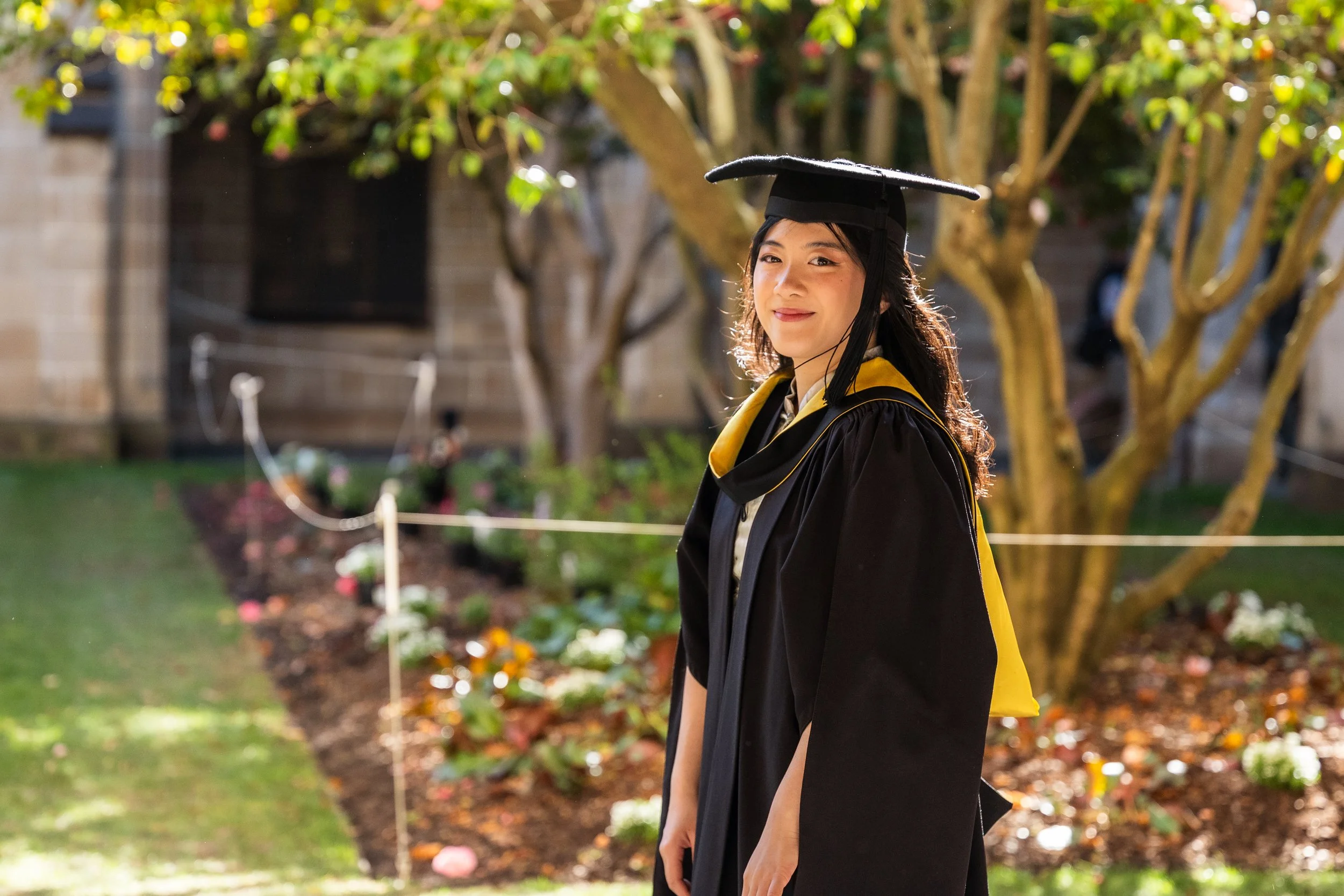 Young woman in graduation gown and cap smiling outdoors during daytime, with trees and flower bed in the background.