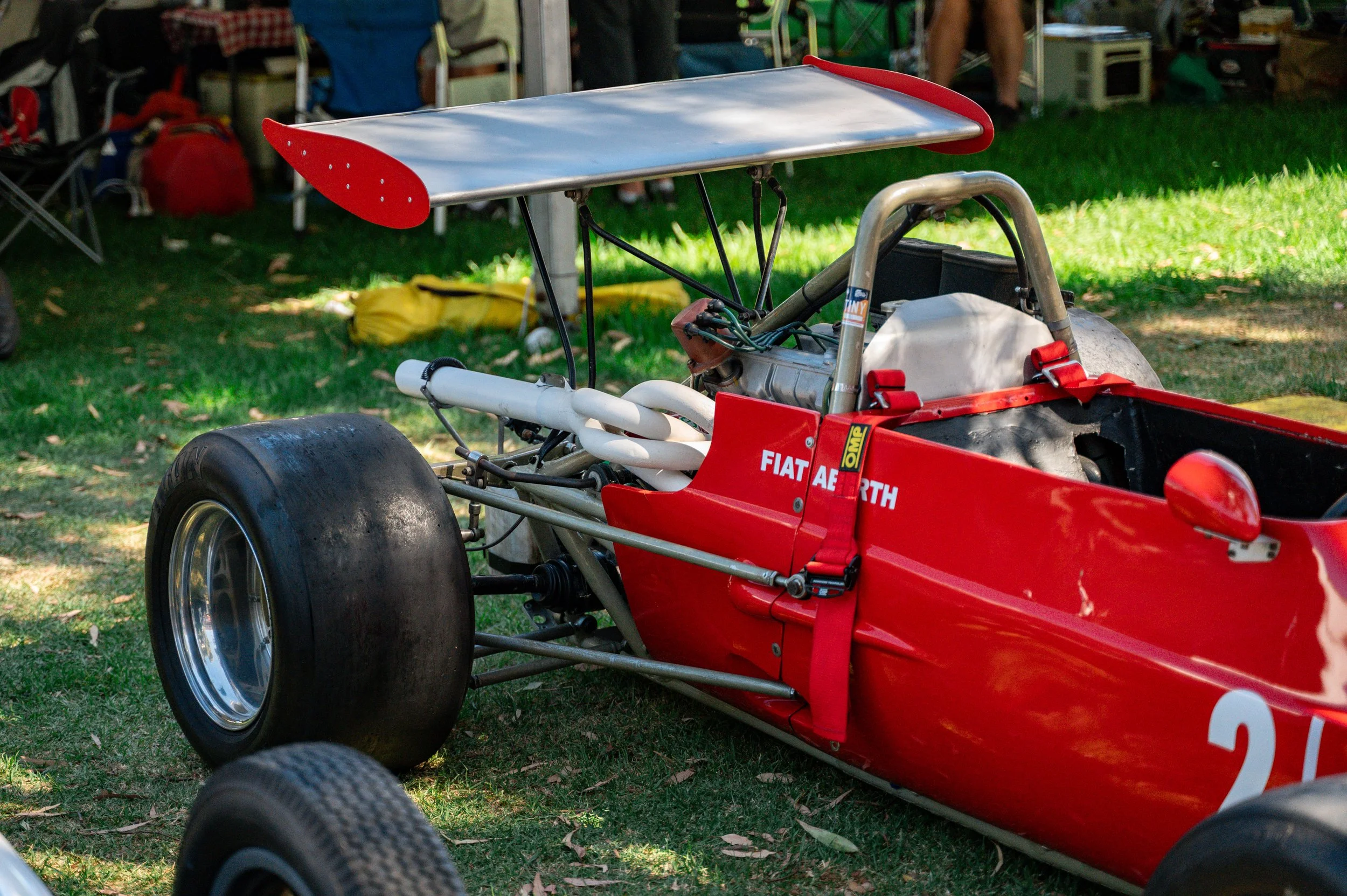 A vintage race car with a red body, large rear tires, and a silver wing on top, parked on grass at a racing event.
