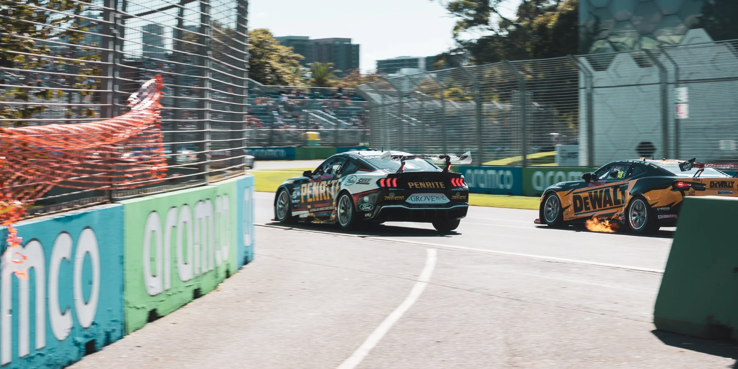 Two race cars collide at a turn during an auto race, with one car emitting flames from its exhaust. The cars are near a barrier with advertisements, and there are spectators in the stands in the background.