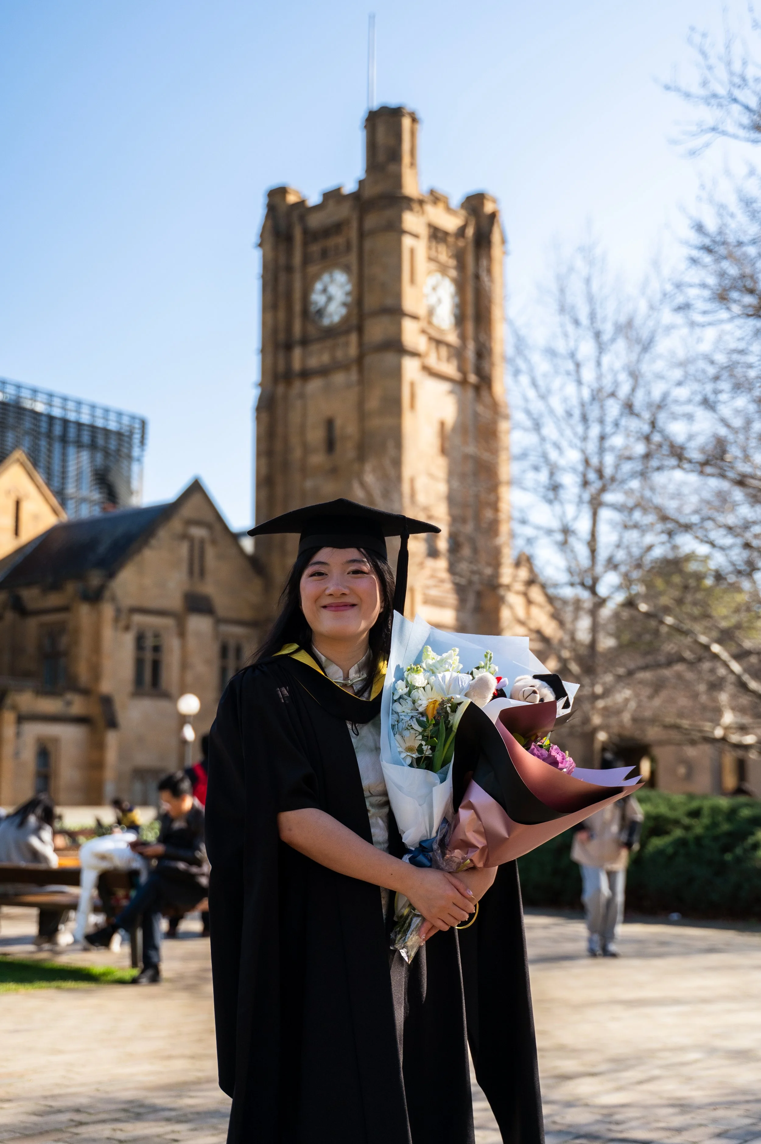 Graduate woman in cap and gown holding a bouquet of flowers, smiling outdoors in front of a historic tower and other people.