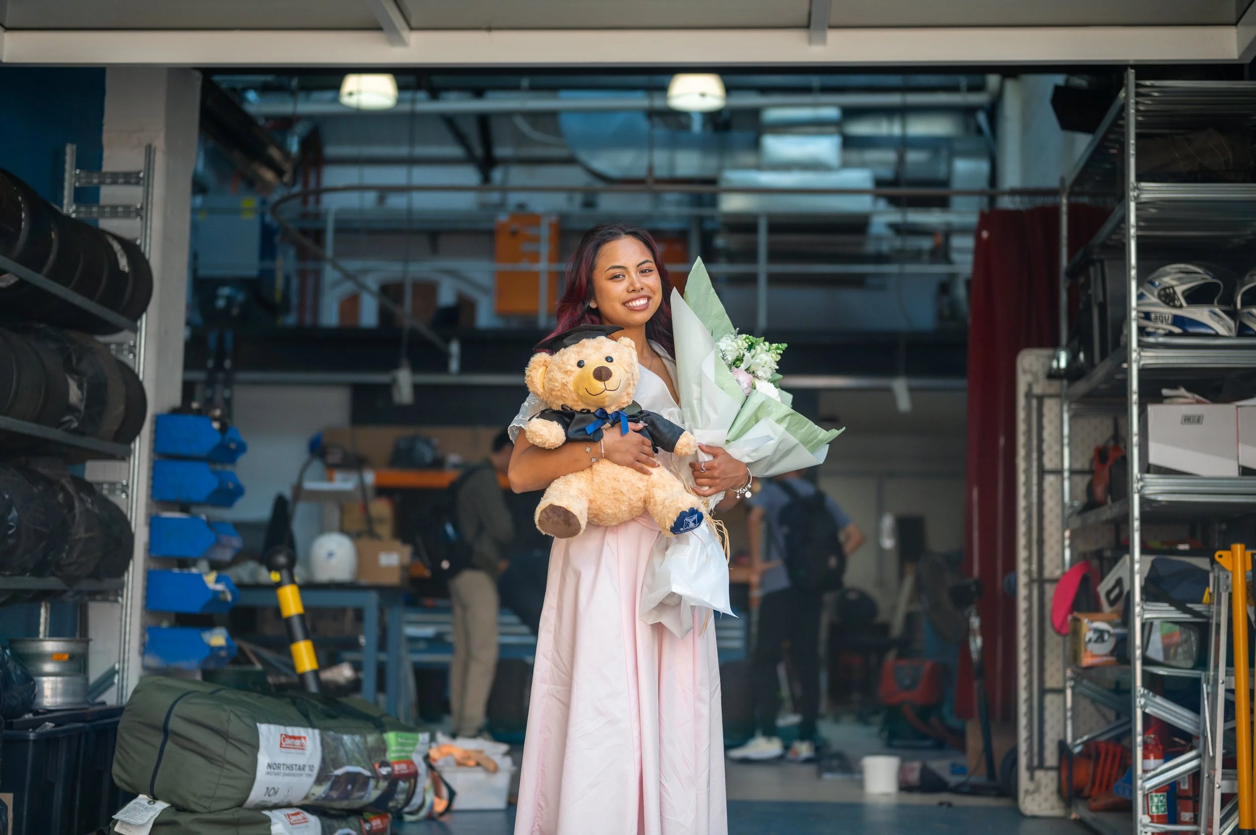 Young woman holding a teddy bear, bouquet of flowers, and smiling in an indoor setting with warehouse shelving and people in the background.