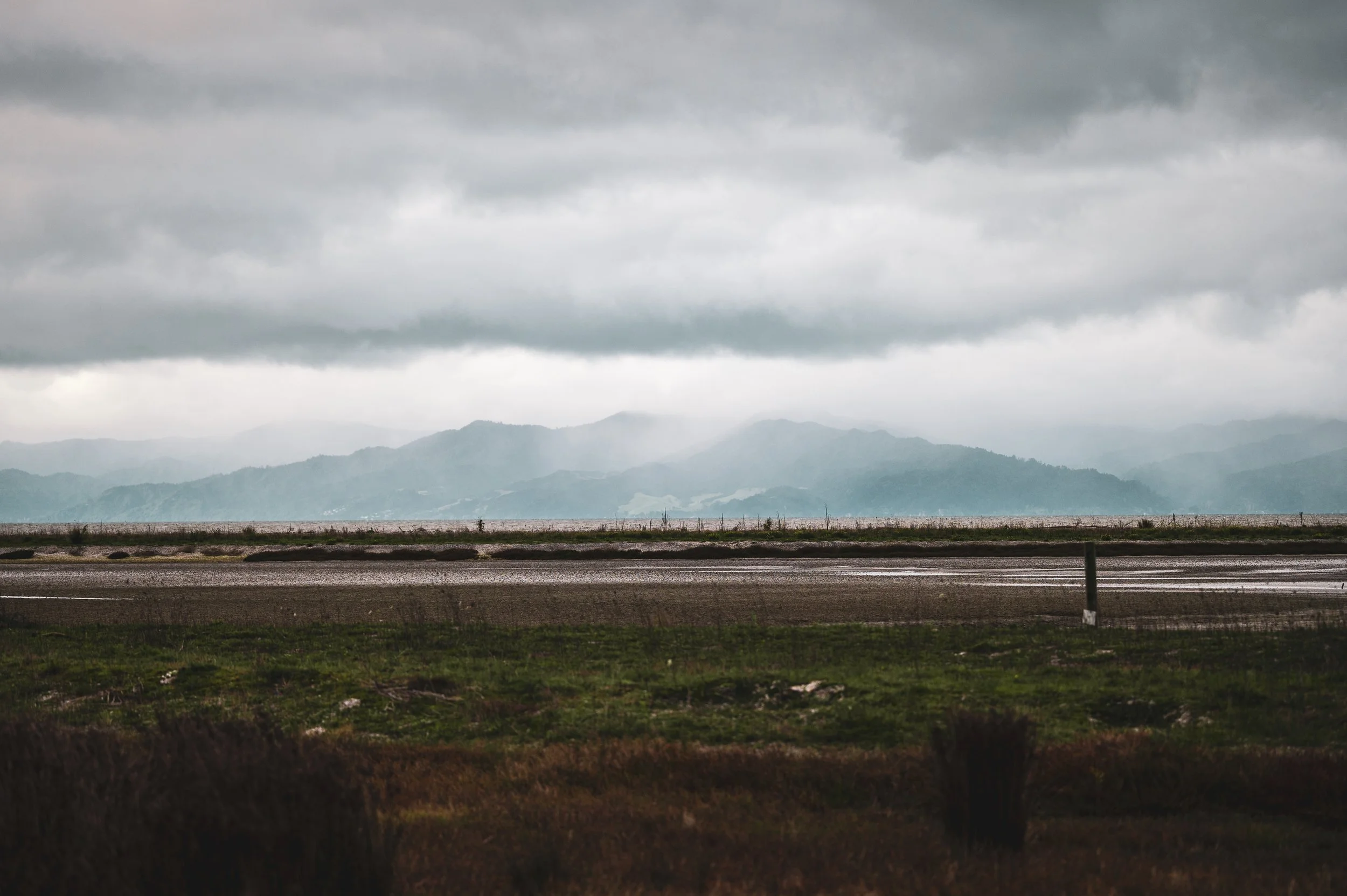 Overcast sky with gray clouds over distant mountains, a narrow road or runway in the foreground with patches of greenery.
